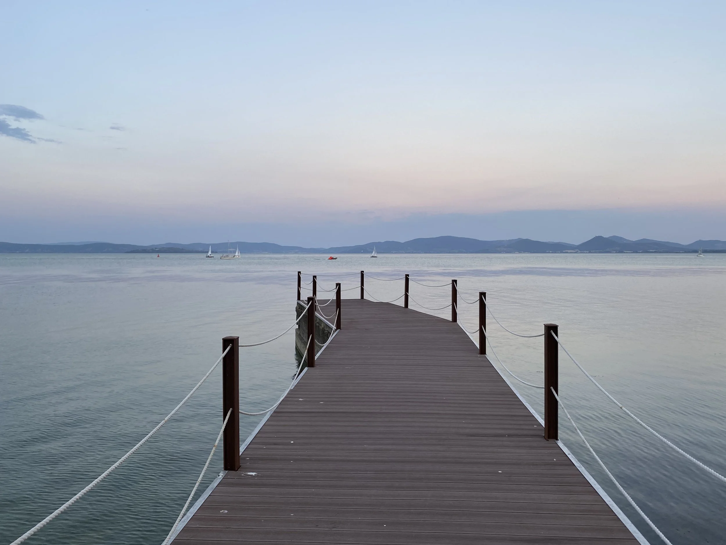 A wooden pier extends out into Lake Trasimeno, with platforms on either side and a rope railing. In the distance, sailboats are sailing, and mountains are visible on the horizon under a pale sky.