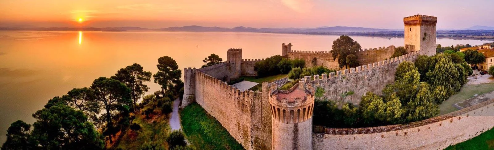 An aerial view of an ancient stone castle with tall towers and fortified walls, situated near a large body of water at sunset, surrounded by lush green trees and a small residential area.