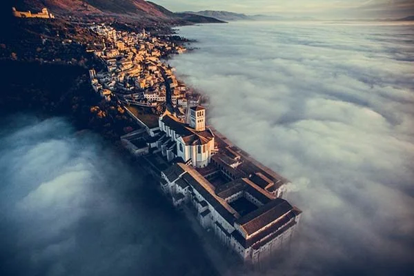 Aerial view of a historic Assisi with a church on top, surrounded by a town, above a thick layer of fog or clouds in a mountainous landscape at sunrise or sunset.