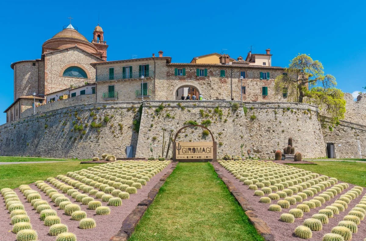 The church at Castiglione del Lago with green shutters and a large dome, under a clear blue sky, with a garden featuring rows of round cacti in the foreground.