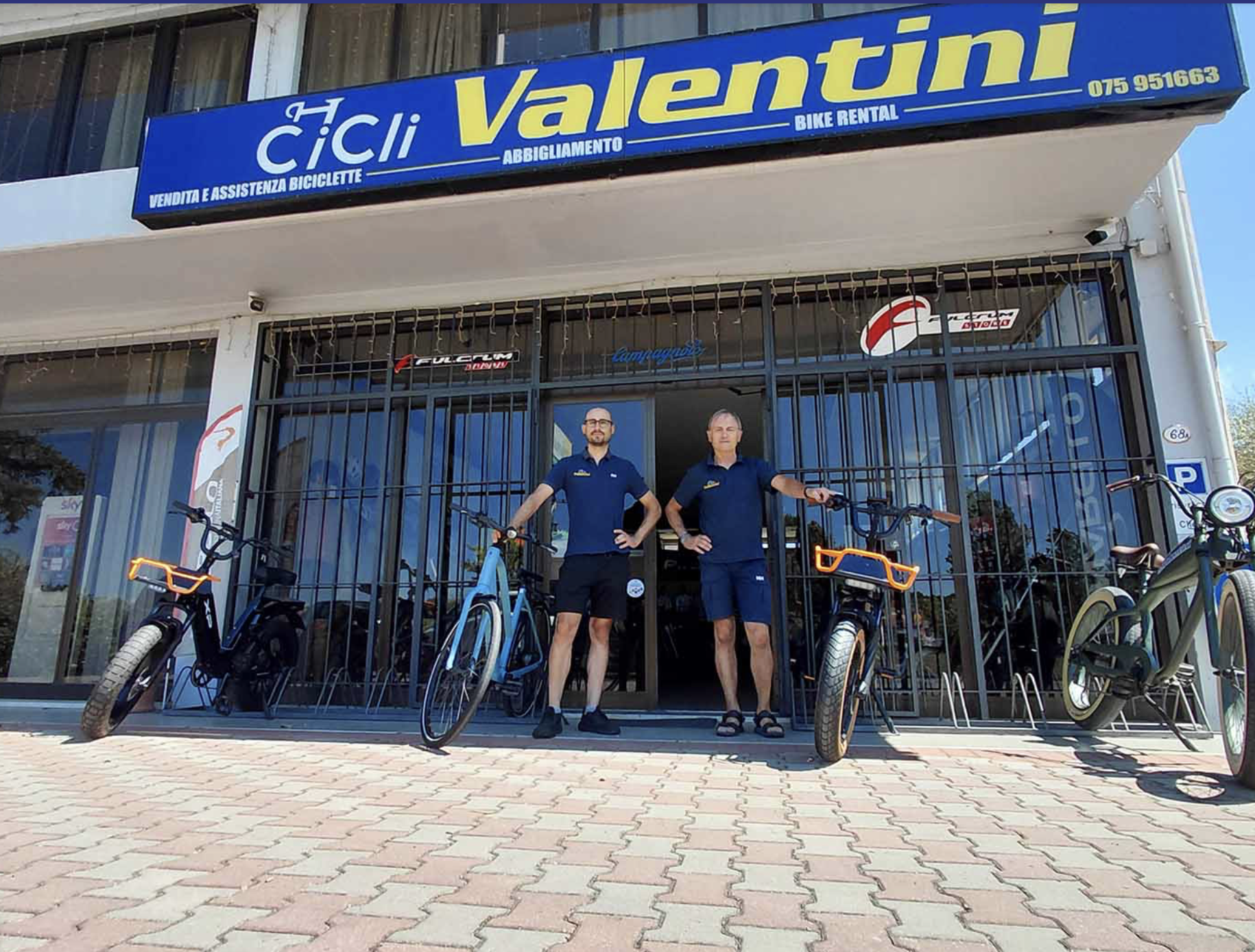 Bike shop with a blue sign reading 'Valentini Bike Rental' and two men standing behind electric bikes outside the shop entrance.