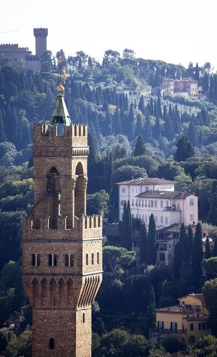 A historic brick tower with battlements and a pointed roof, set against a hillside with trees and residential buildings, under a clear sky in Florence.