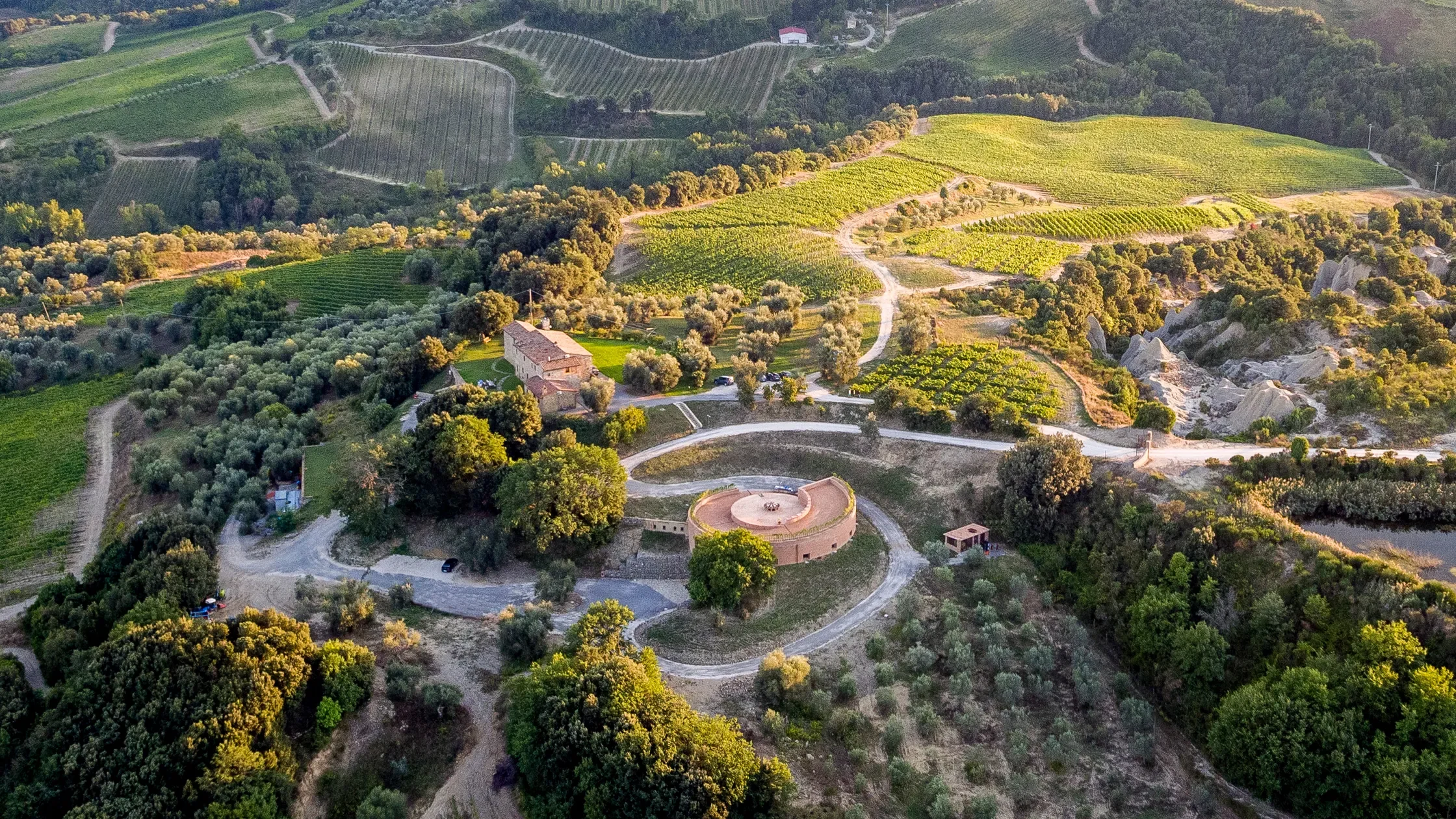 Aerial view of a hilly, lush green landscape with vineyards, trees, and scattered buildings, including a circular structure and a winding dirt road.