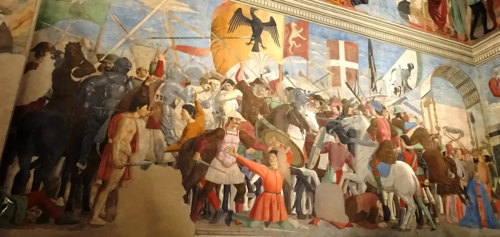 Fresco by Piero Della Francesca depicting a medieval battle scene with knights on horseback, soldiers, banners, and flags, in Arezzo.