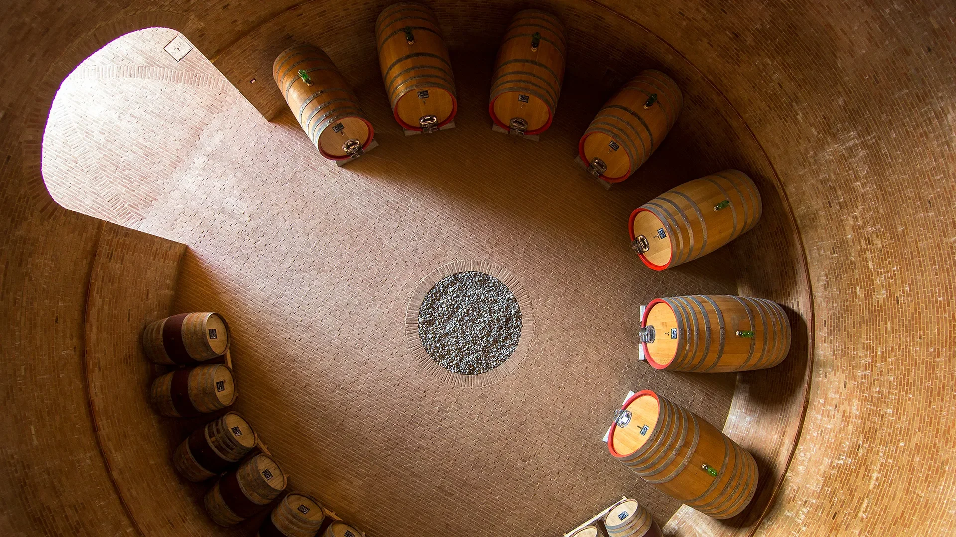 A wine cellar with a circular brick ceiling and walls. Several wine barrels are stacked vertically on the wall and placed on the floor, with a round window or decorative opening in the ceiling.