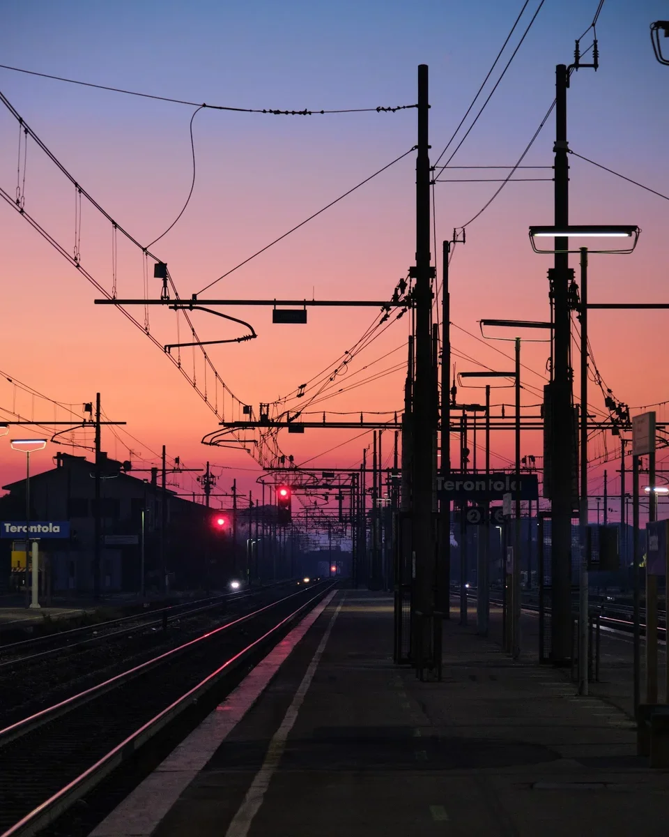 Silhouette of train station platform in Terontola at sunset with overhead power lines and tracks in the foreground, colorful sky with pink, purple, and blue hues in the background.