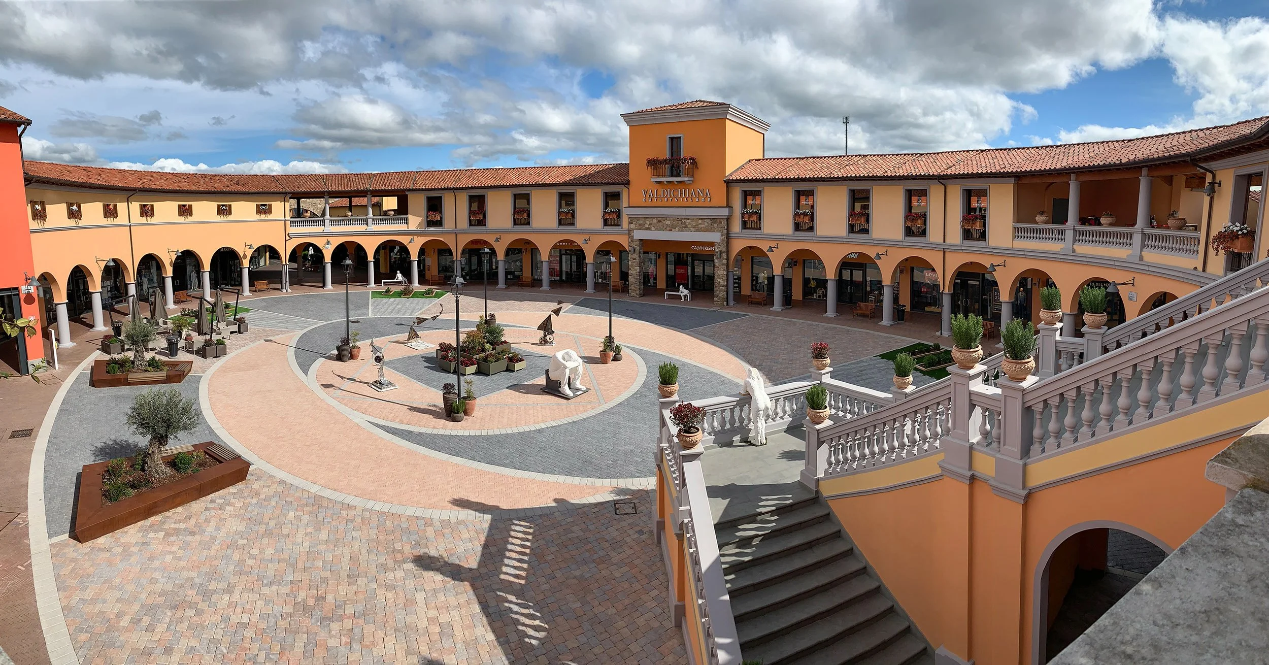View of a Mediterranean-style shopping plaza with a central courtyard, decorative sculptures, potted plants, and storefronts with arched windows and balconies.