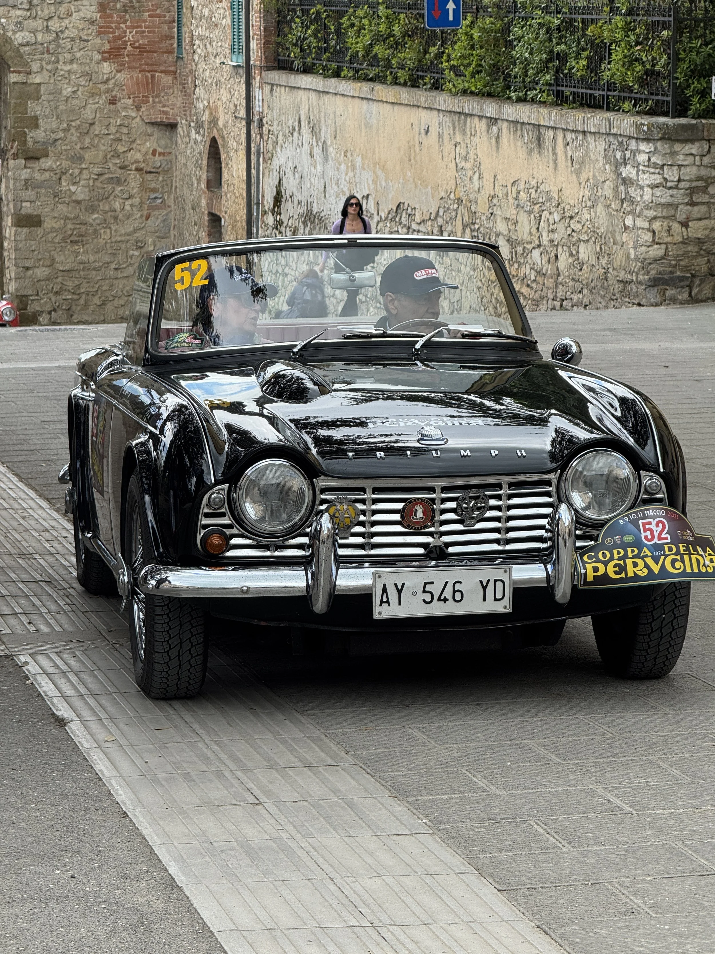 A black vintage Triumph convertible car with the number 52 on its windshield, parked on a street with a brick and stone wall in the background, in Castiglione Del Lago.