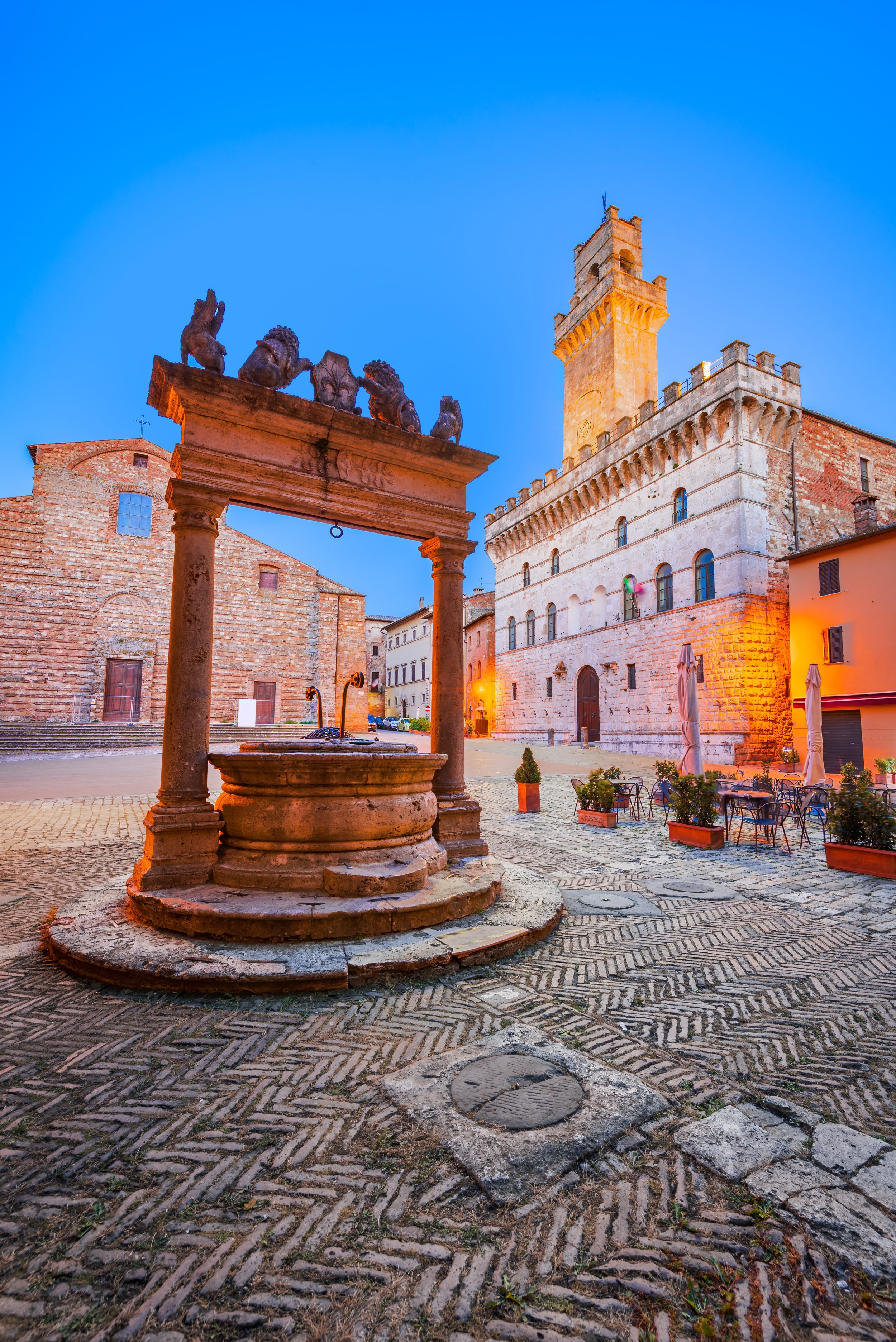 Historic Montepulciano town square at dusk with a stone fountain in the foreground, ancient brick buildings, and a church tower illuminated against a blue evening sky.