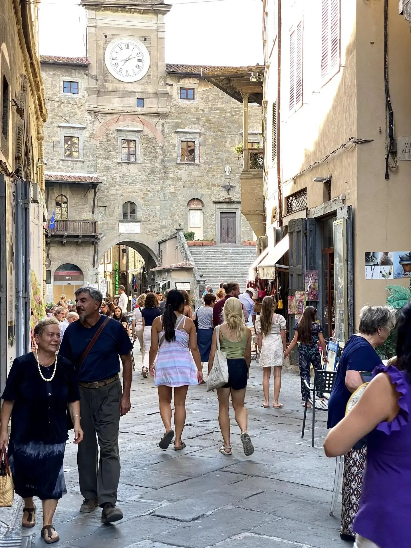 Crowded street scene in Cortona with people walking and shopping near historic stone buildings with clock tower in the background.