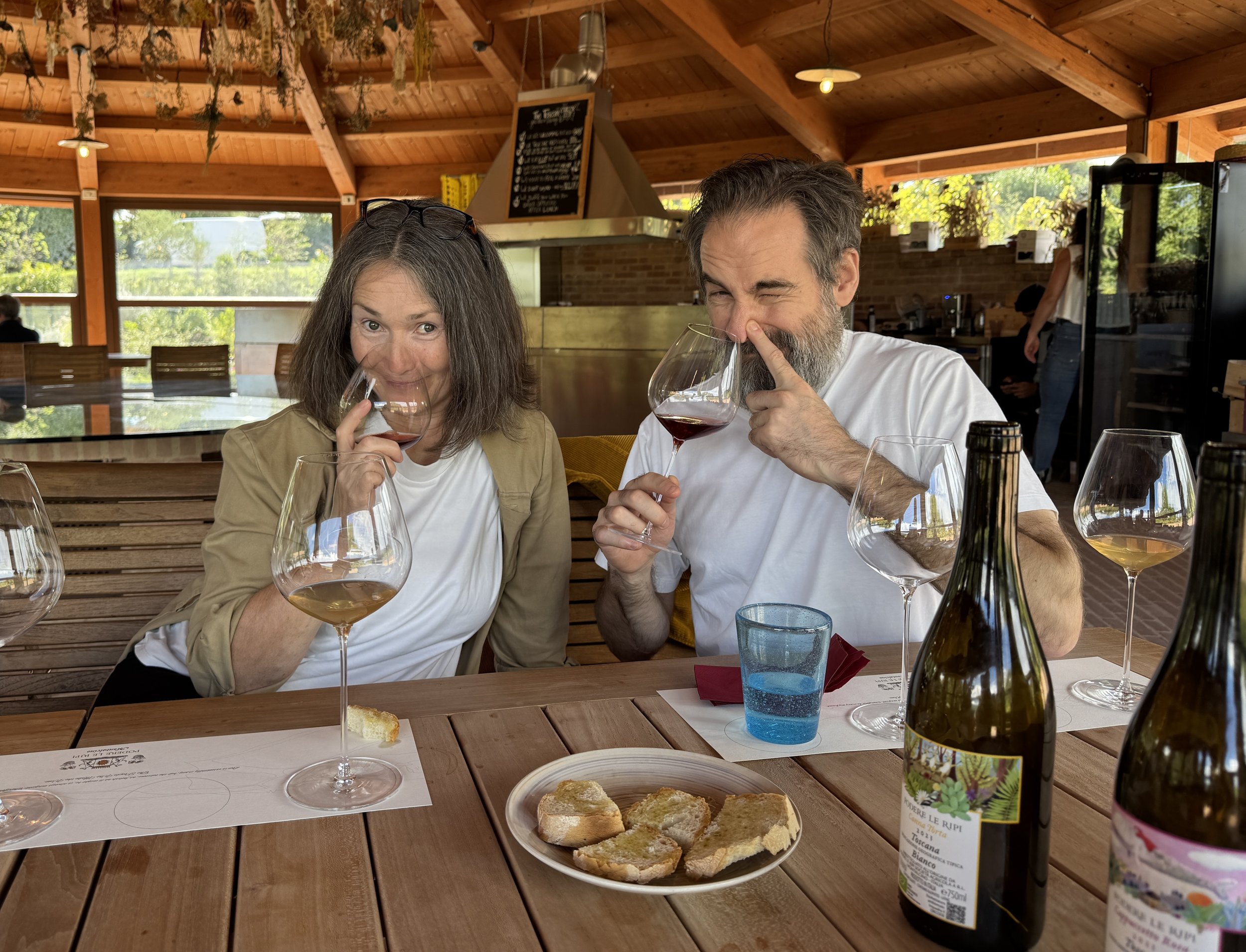 Two people enjoying wine at a wooden table inside a restaurant with large windows and natural light. There are wine bottles and snack bread on the table.