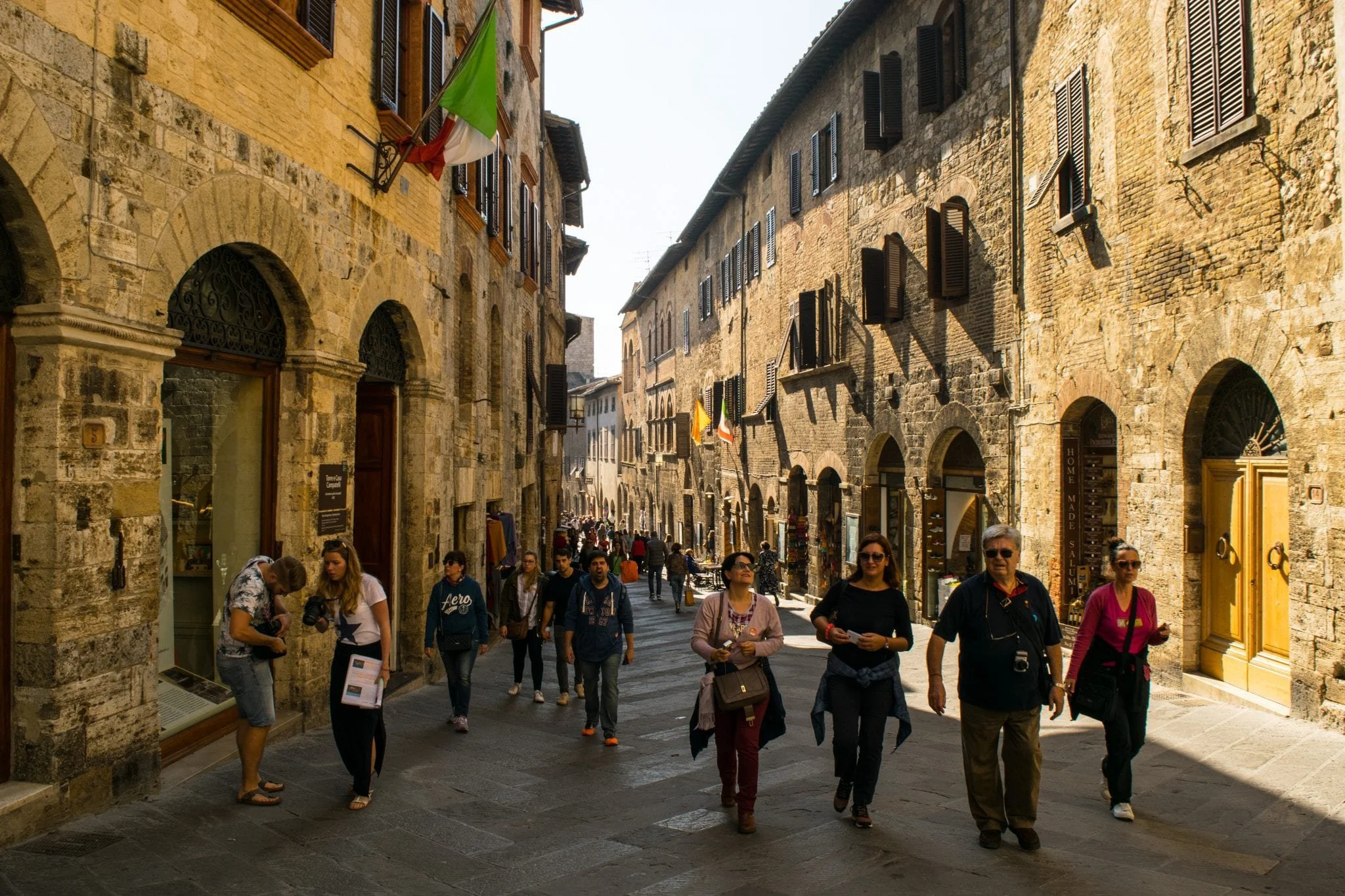 People walking along a narrow street in Arezzo with stone buildings, some with black window shutters, and flags hanging from the buildings.
