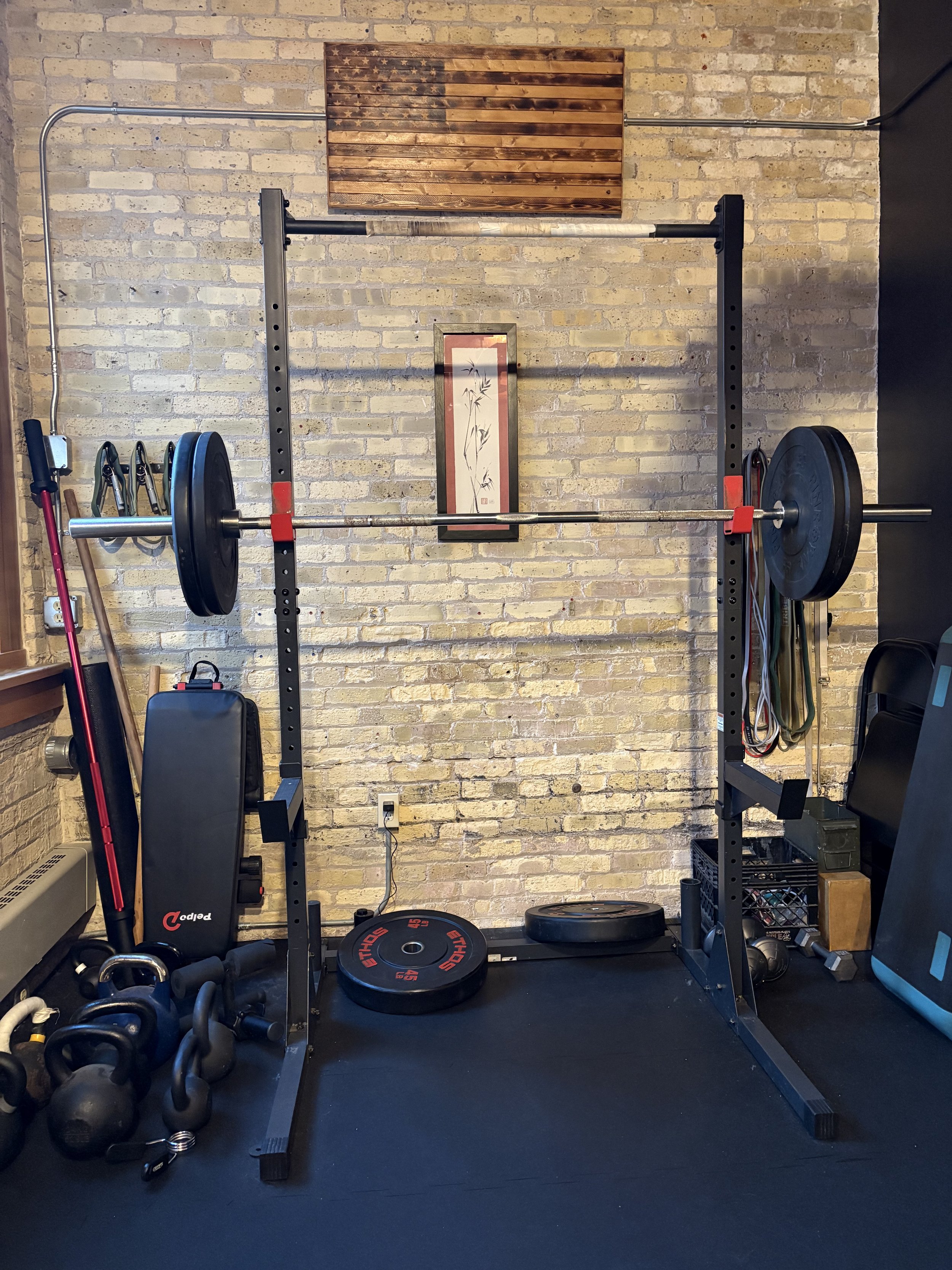A home gym with a steel squat rack, adjustable bench, bumper plates, kettlebells, foam roller,  a barbell, and resistance bands against a brick wall with a framed Asian-style artwork and wooden american flag