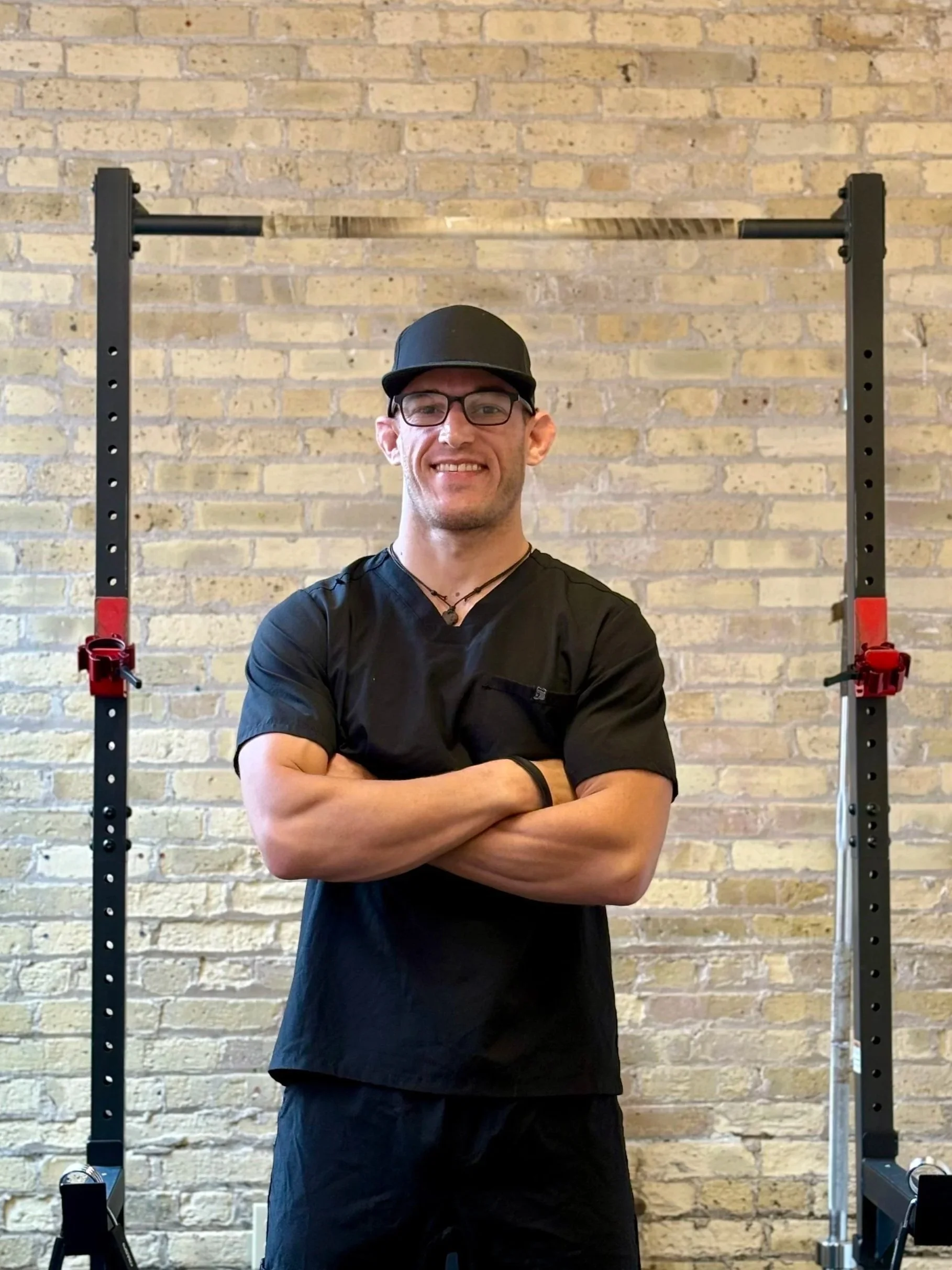 A man wearing glasses and a black cap smiling with his arms crossed in front of a brick wall, standing in front of a black metal structure.