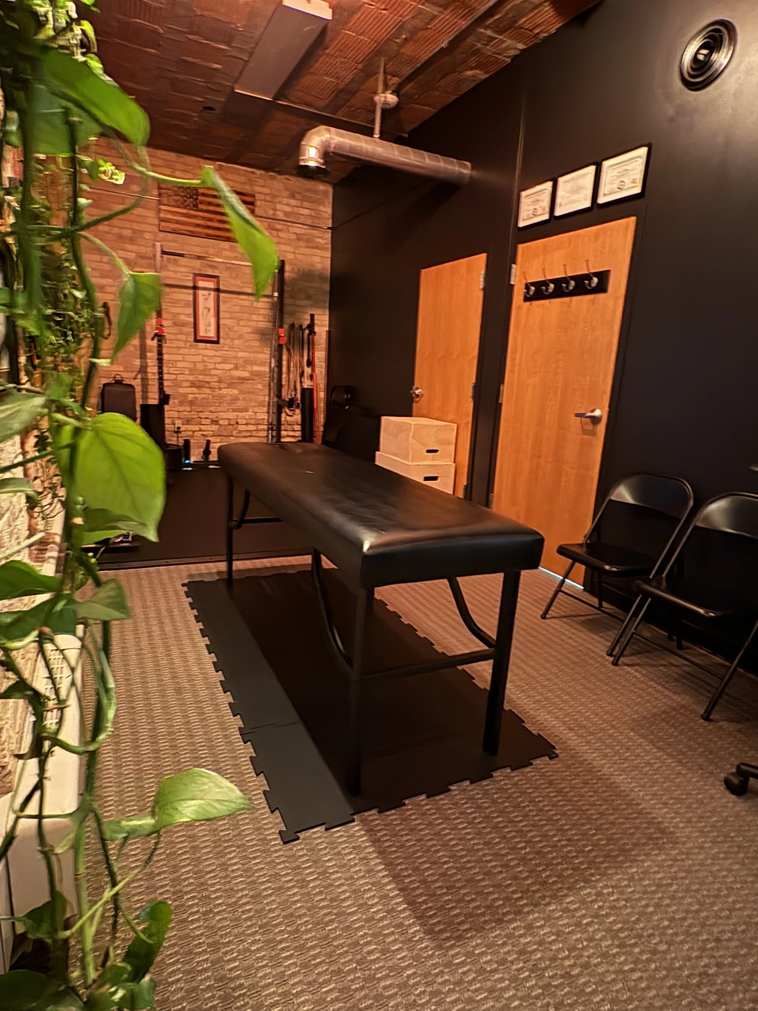 A room with a brick wall, black ceiling with exposed ductwork, wooden doors, a black massage table, black chairs, and framed certificates or artwork on the wall. There are plants on the left side and a beige rug on the floor.