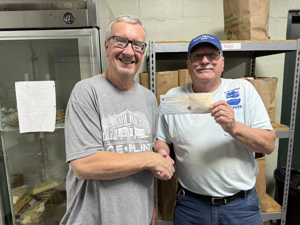 Two smiling men shaking hands, one holding a check, in a storage room with shelves and paper bags.