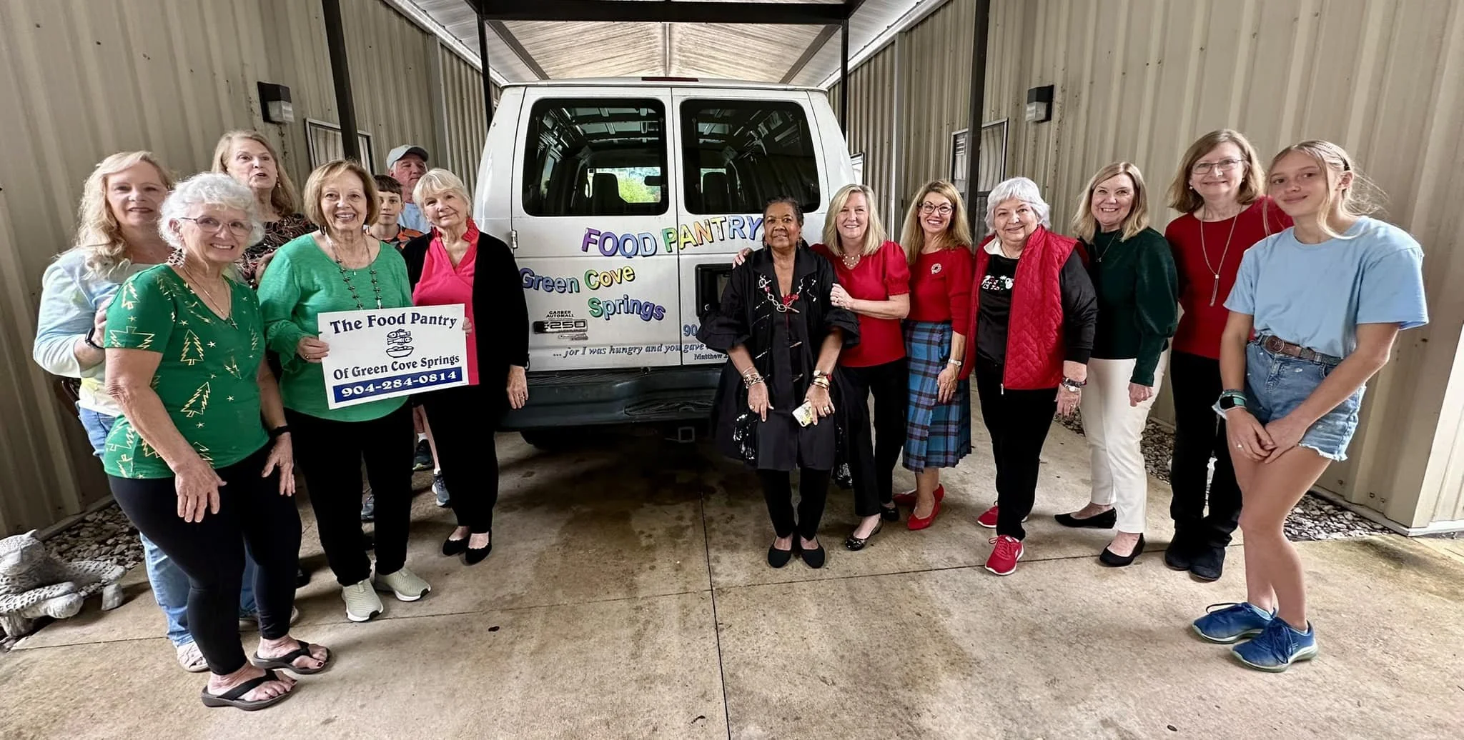 A group of women standing in front of a white Food Pantry vehicle with colorful signs for Green Cove Springs Food Pantry. Some women are holding a sign with the pantry's name and phone number, smiling for the camera.