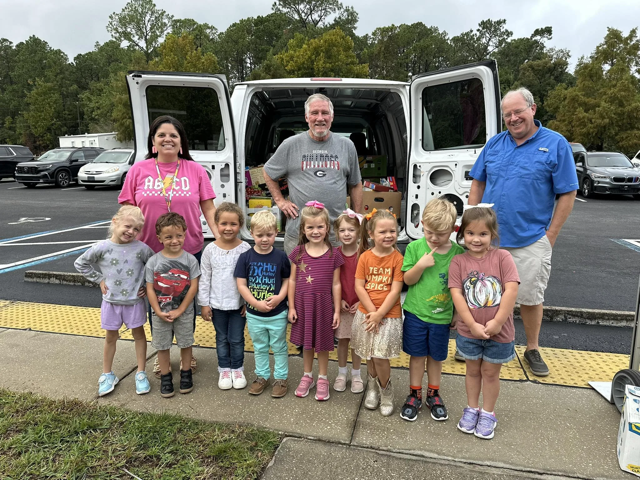 Group of children and adults standing in front of a white van with open doors, with donation boxes and supplies inside, in a parking lot surrounded by trees.