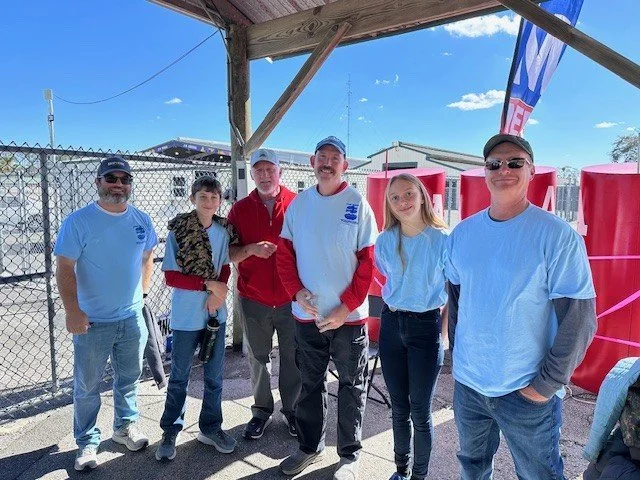 Group of six people standing outdoors under a wooden shelter, smiling for the photo. They are dressed casually, with some wearing t-shirts and baseball caps. Behind them are industrial buildings and a chain-link fence, with a blue sky and clouds overhead.