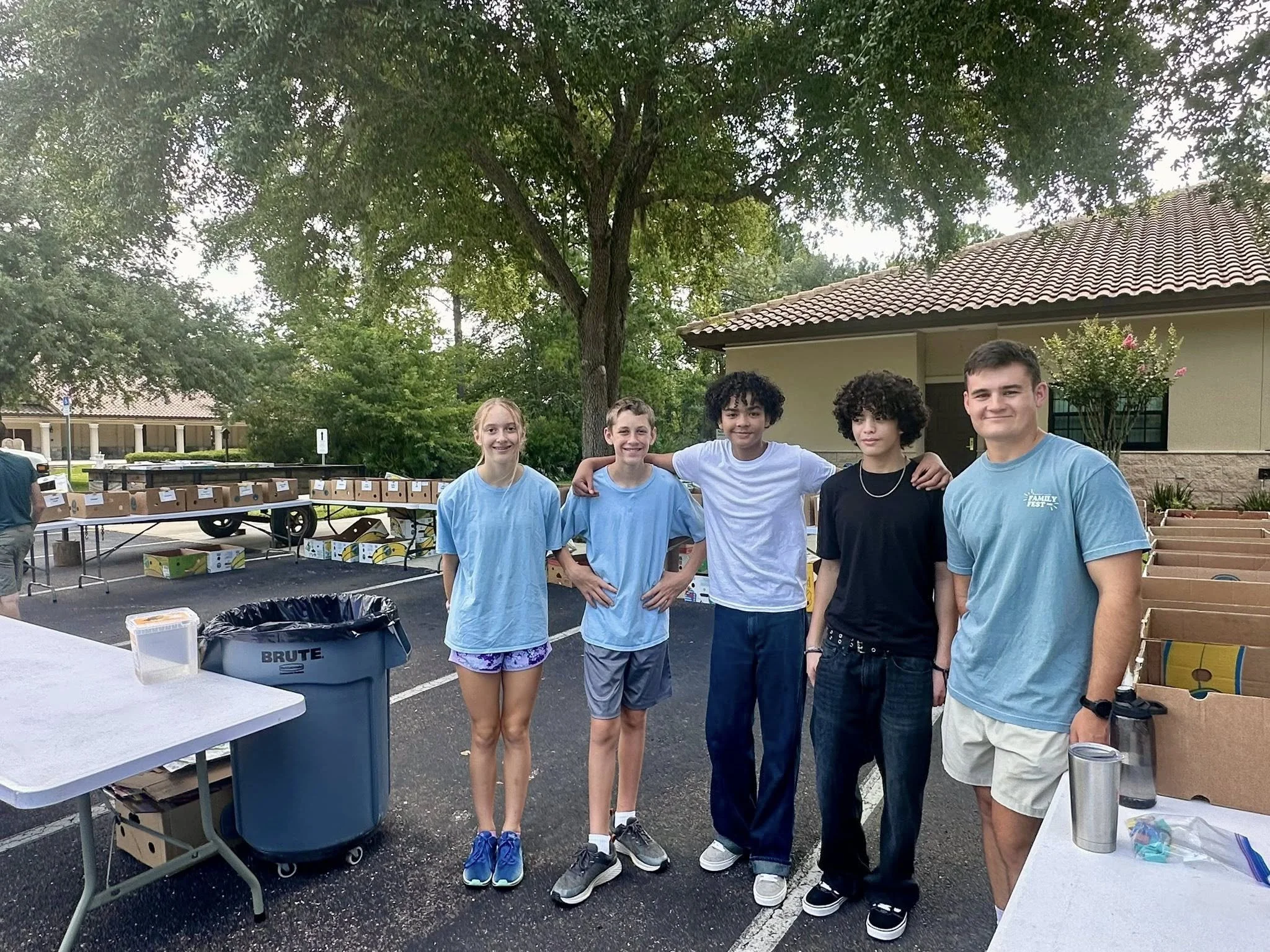 Group of five teenagers standing together outdoors near tables with boxes, a blue trash bin, and a building with a tiled roof, trees, and green foliage in the background.