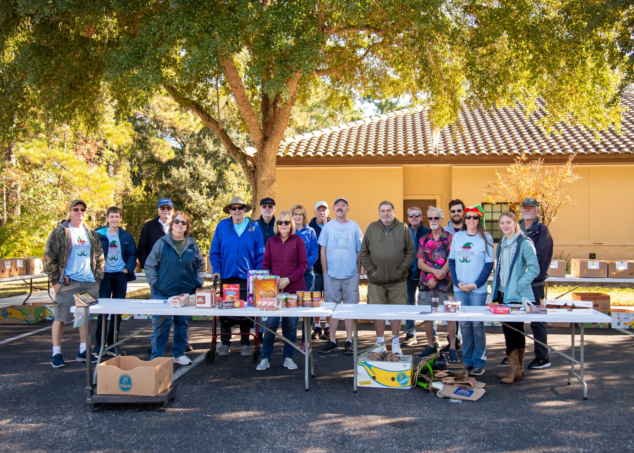 Group of people standing behind tables with food donations outdoors under a large tree.
