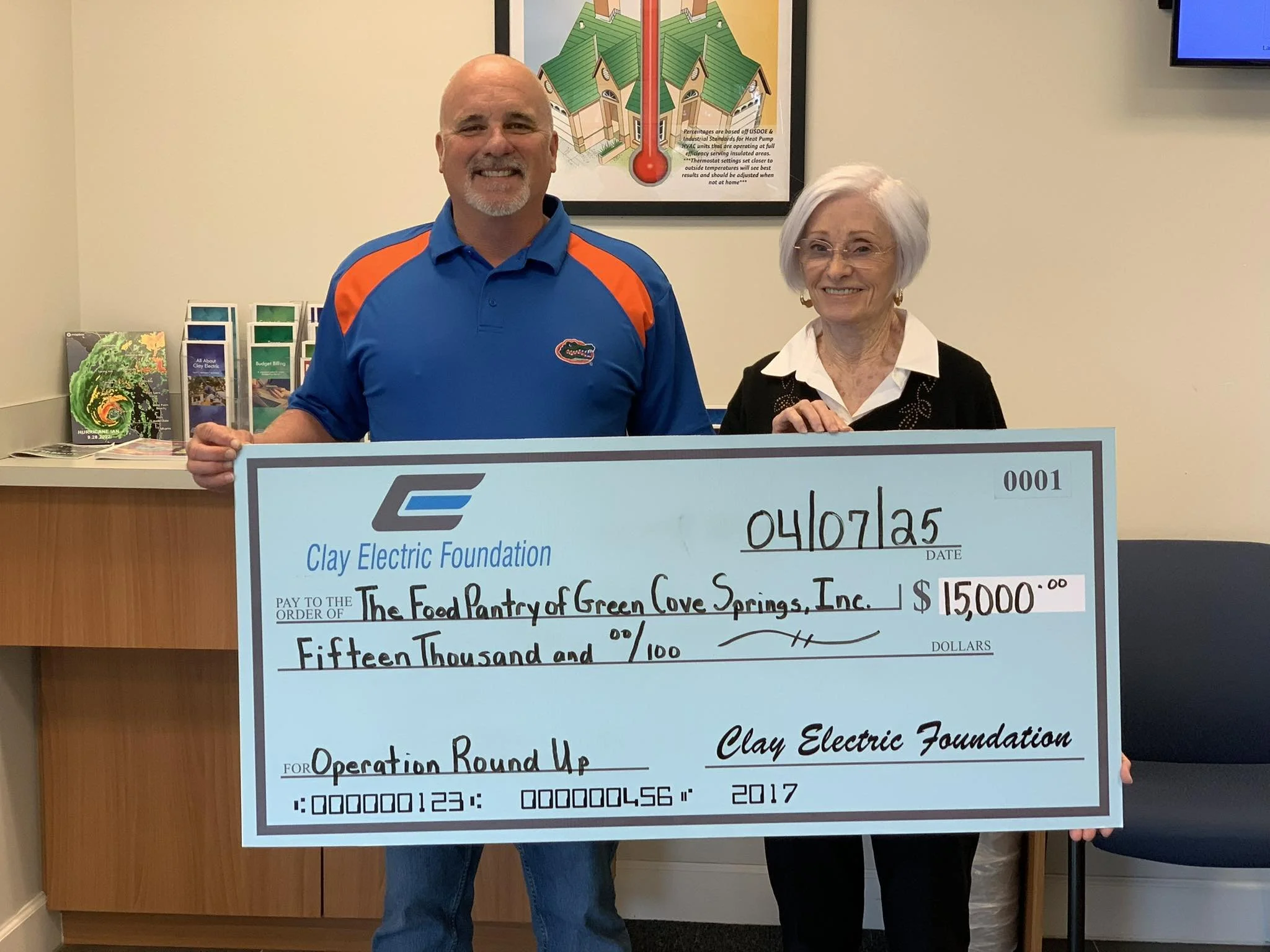 A man and a woman holding a large check from the Clay Electric Foundation for $15,000 made out to the Food Pantry of Green Cove Springs Inc. The man is wearing a blue shirt with orange accents, and the woman has white hair, glasses, and is wearing a white blouse with a black sweater. Behind them is a counter with brochures, a colorful poster about weather safety, and a blue chair.
