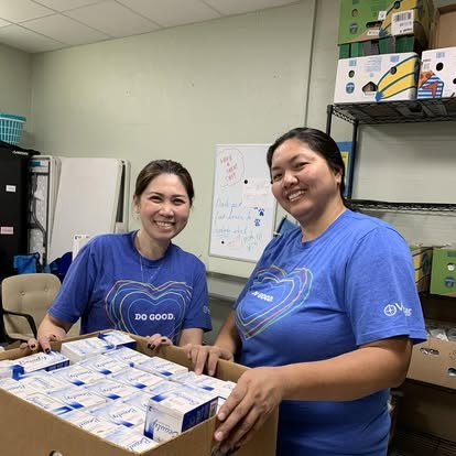 Two women in blue shirts smiling at the camera, packing or organizing boxes of medical supplies in a storage room.