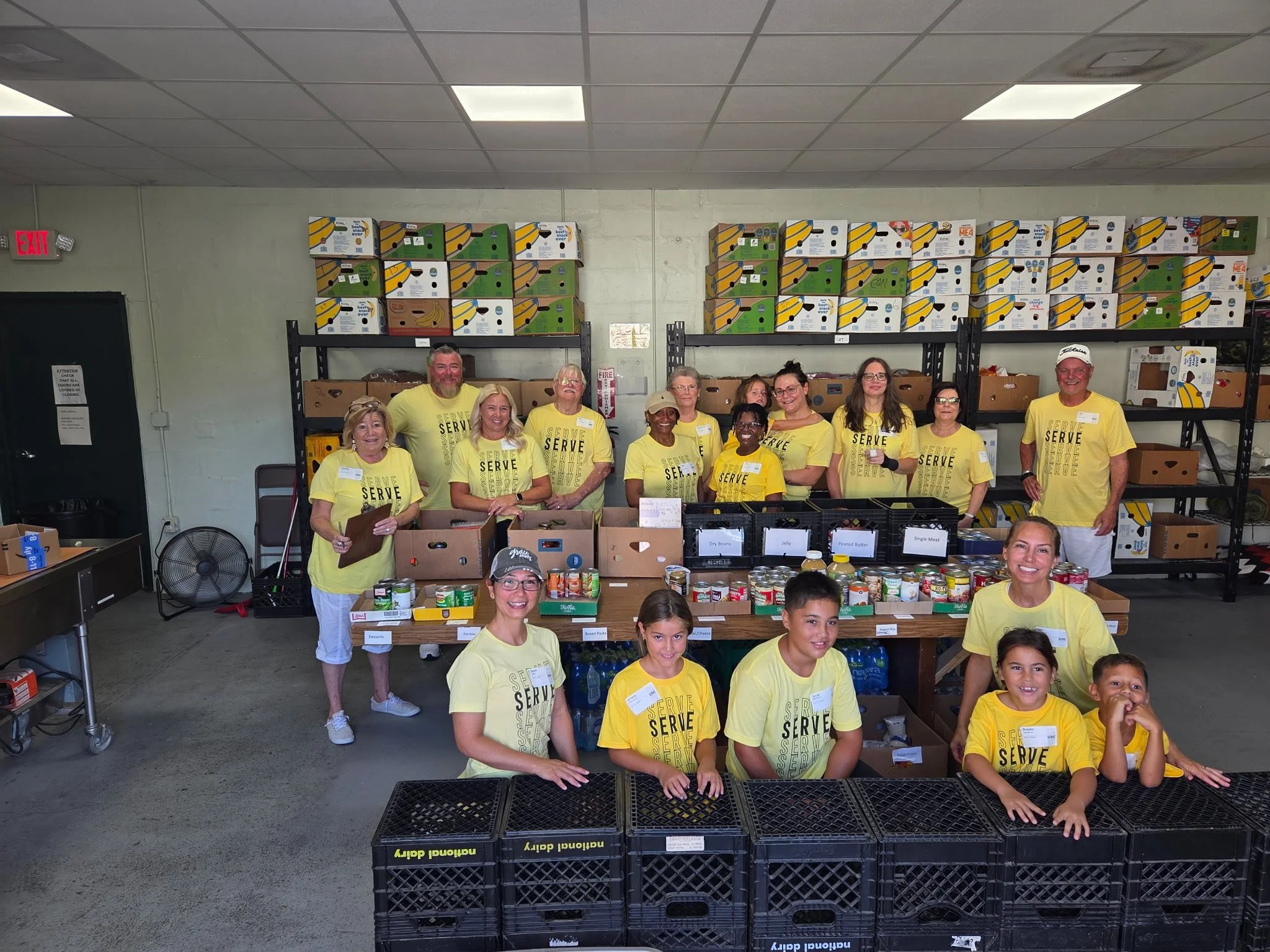 Group of volunteers in yellow shirts working in a storage room with boxes of food and supplies for a food drive or community service event.