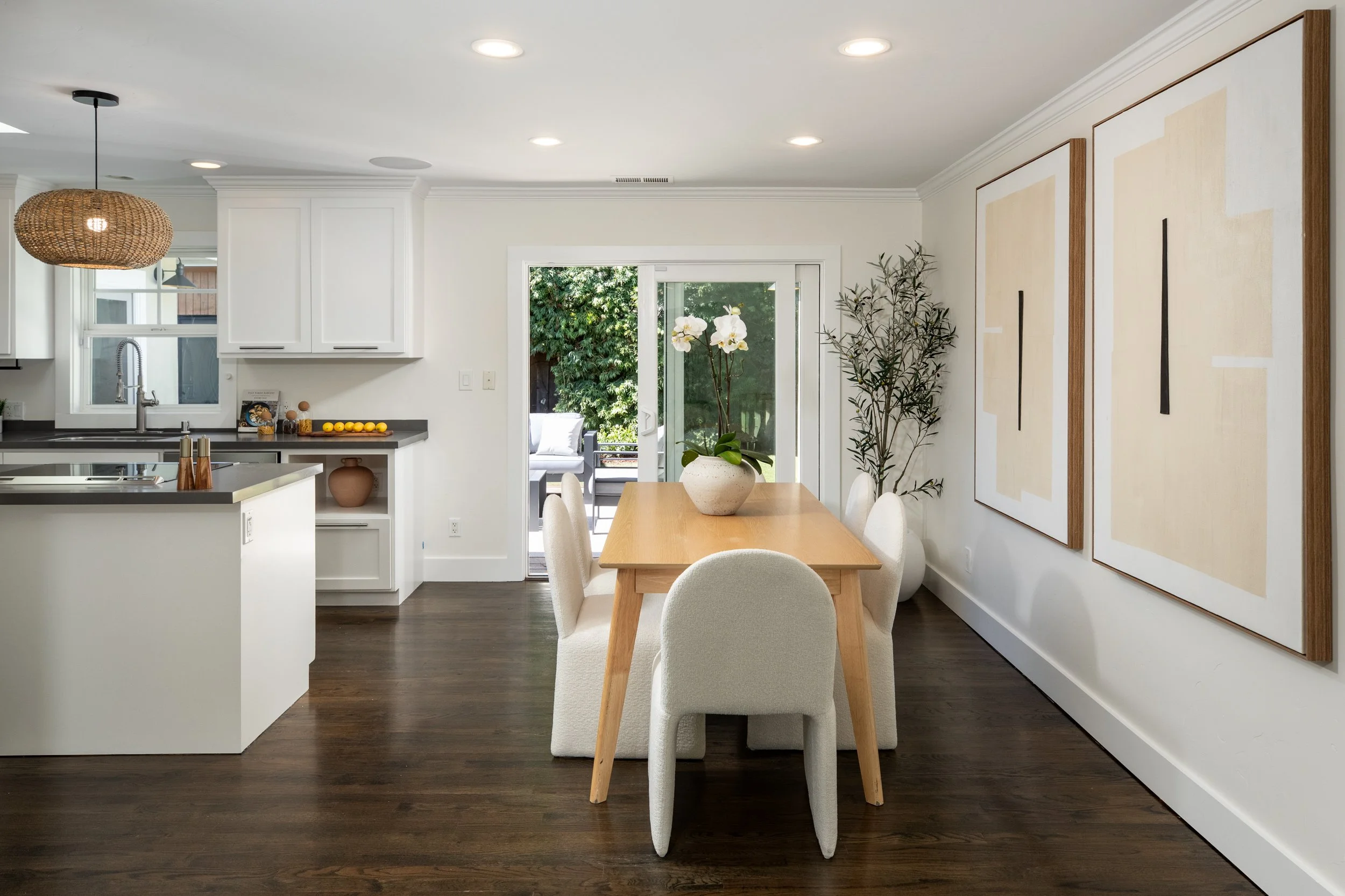 A dining room with a wooden table and six white upholstered chairs. A large pot with white orchids on the table. A garden with green trees visible through a sliding glass door. White walls with large abstract paintings. Recessed ceiling lights and a 