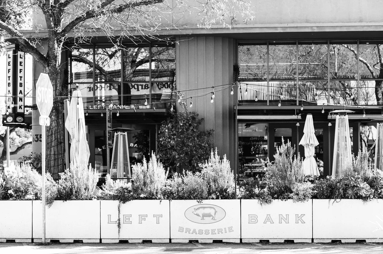 Exterior of a brasserie called 'Left Bank,' with outdoor seating, umbrellas, heaters, plants, and string lights, black and white photo.