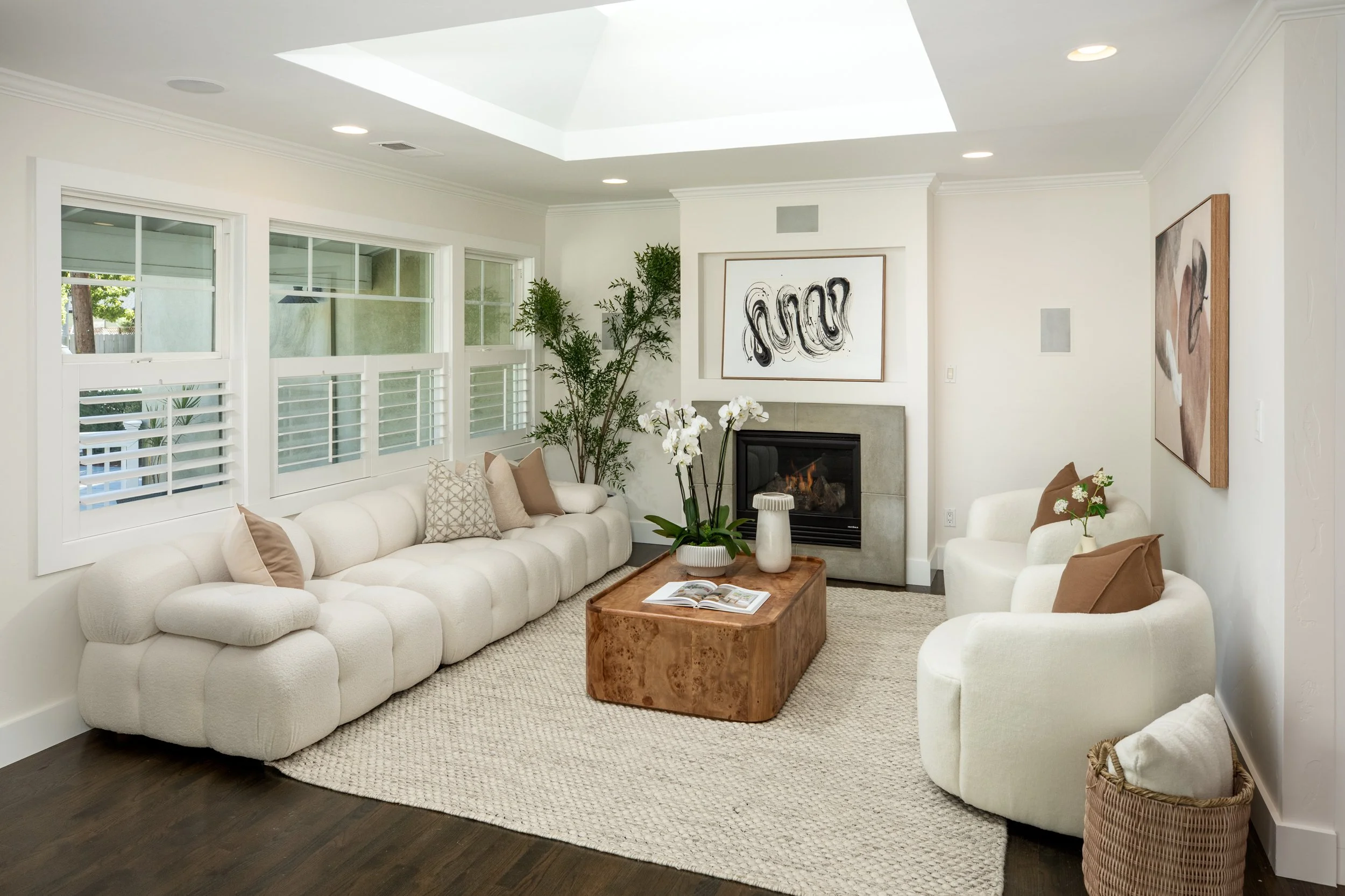 Living room with white sectional sofa, wooden coffee table, fireplace, large windows with blinds, artwork on walls, and indoor plants.