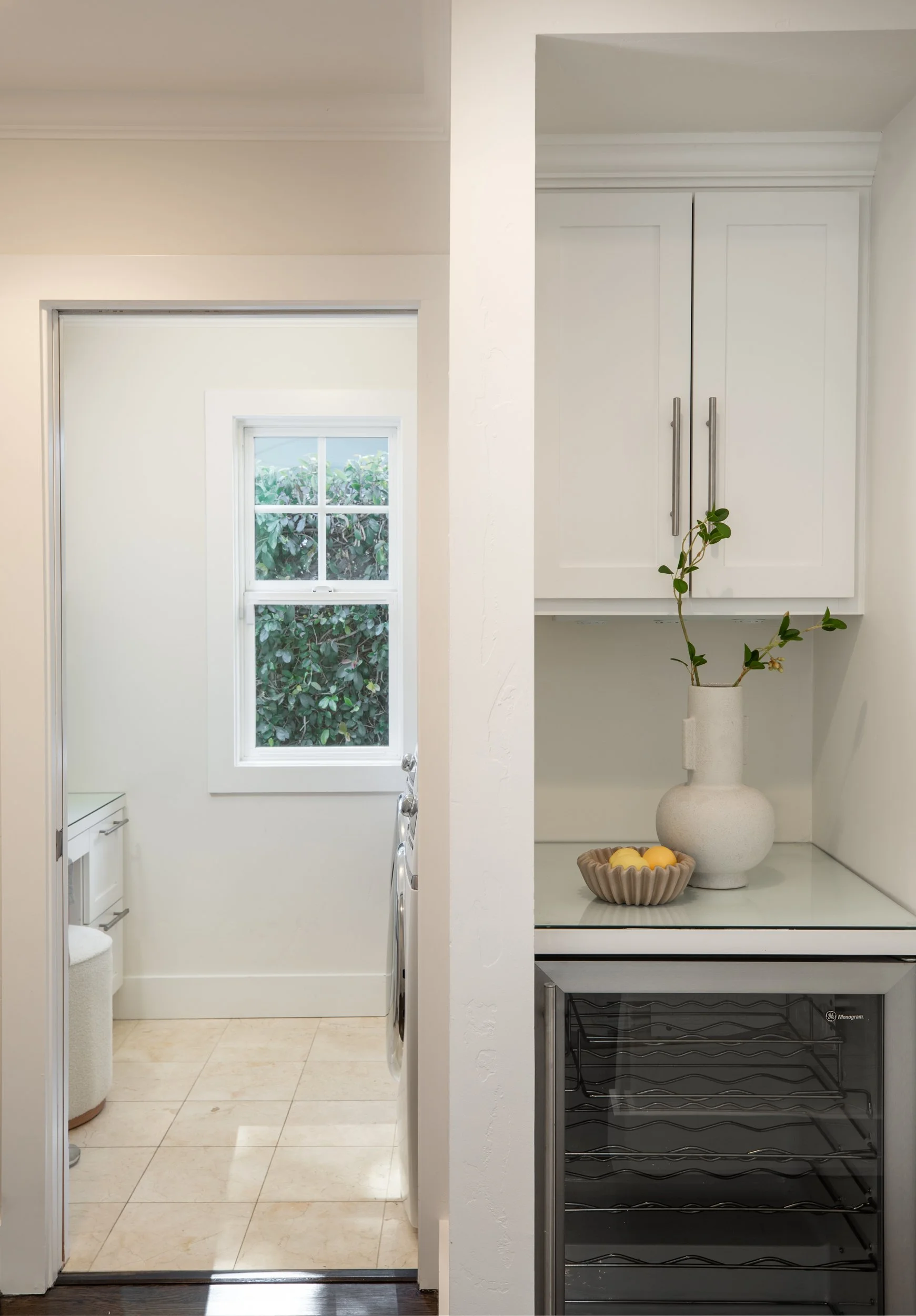 A view from a kitchen into a laundry and back door area with a window showing greenery outside. On the right, a white cabinet with a potted plant and a bowl of lemons, while on the left, a washing machine and beige tiled floor.