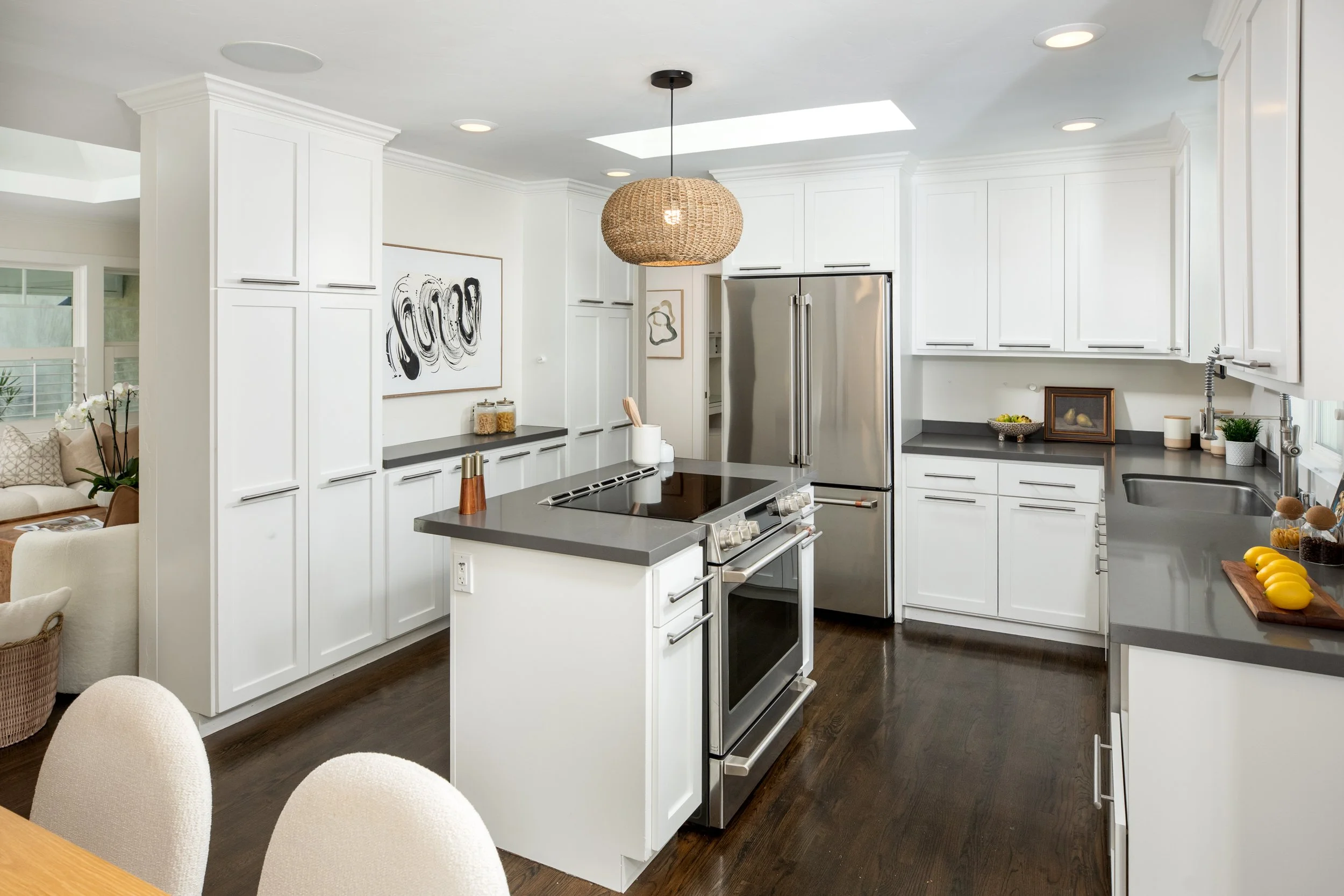 A modern kitchen with white cabinets, black countertops, and a stainless steel refrigerator. There is a kitchen island with a black stovetop, and various kitchen items and art decorations on the counters. The kitchen has dark wooden floors and a hang