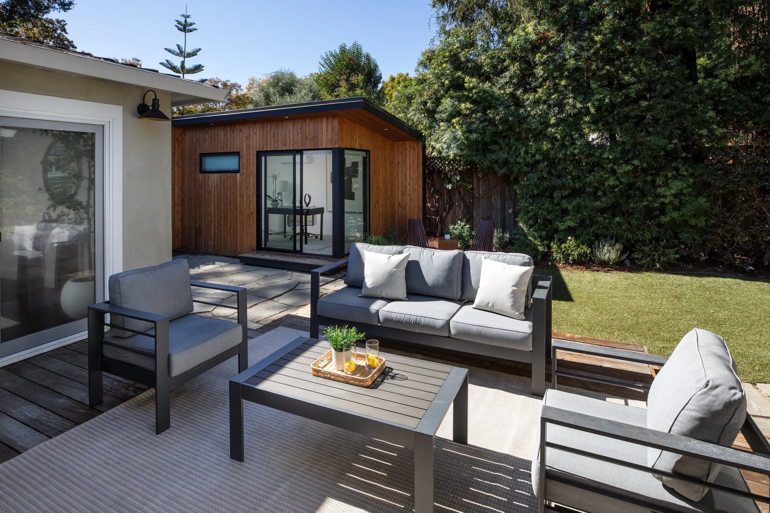 Outdoor patio with gray seating, a low coffee table with drinks, a wooden deck, and a small wooden outdoor structure with a glass door. Lush greenery with trees and shrubs surrounds the area.