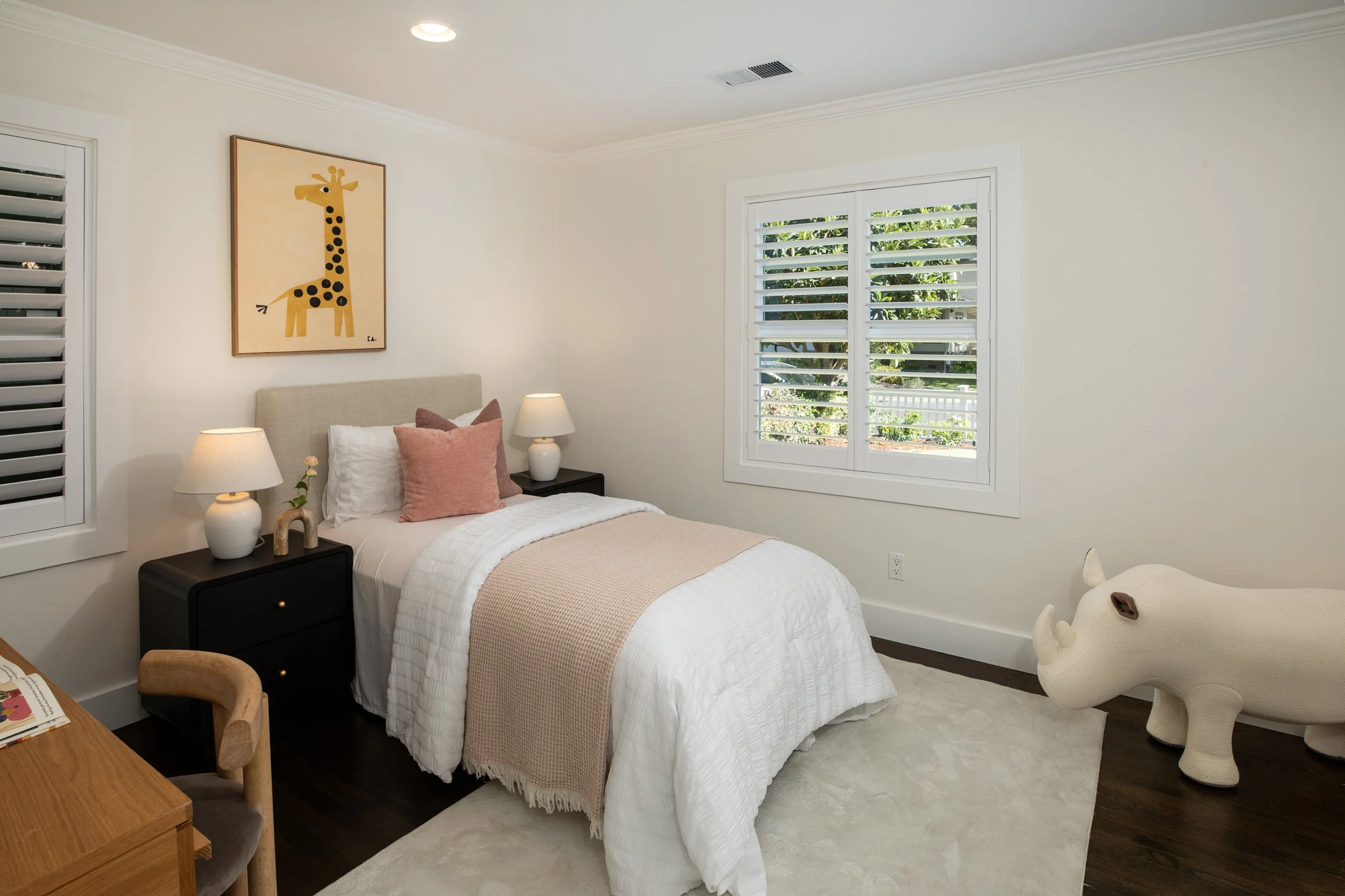 A cozy bedroom with white walls and a bed covered in white linens, beige blanket, and pink pillows. There are two white lamps on black nightstands, a giraffe-themed artwork on the wall, a white louvered window with greenery outside, a cream-colored p
