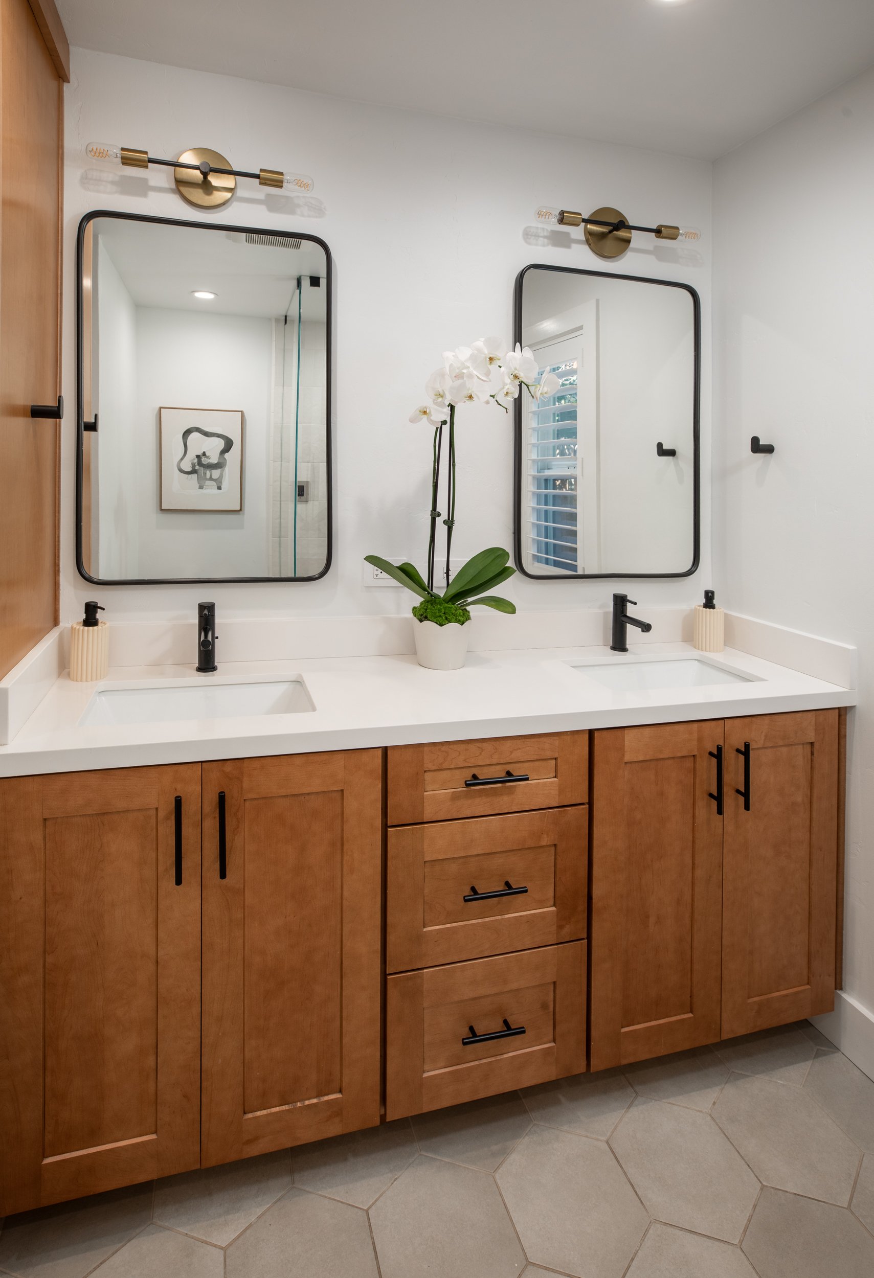 Bathroom vanity with two sinks, black faucets, a white countertop, wooden cabinet with black handles, two mirrors, wall-mounted light fixtures, a potted orchid plant, and decorative soap dispensers.