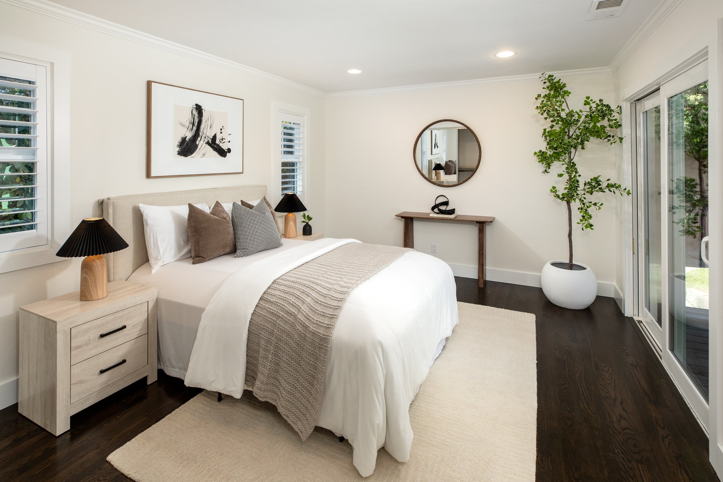A cozy bedroom with a large bed decorated with white and gray pillows, a beige throw blanket, and two black-lamp bedside tables. The room features modern art, a round mirror, a potted tree near sliding glass doors, and a wooden console table.