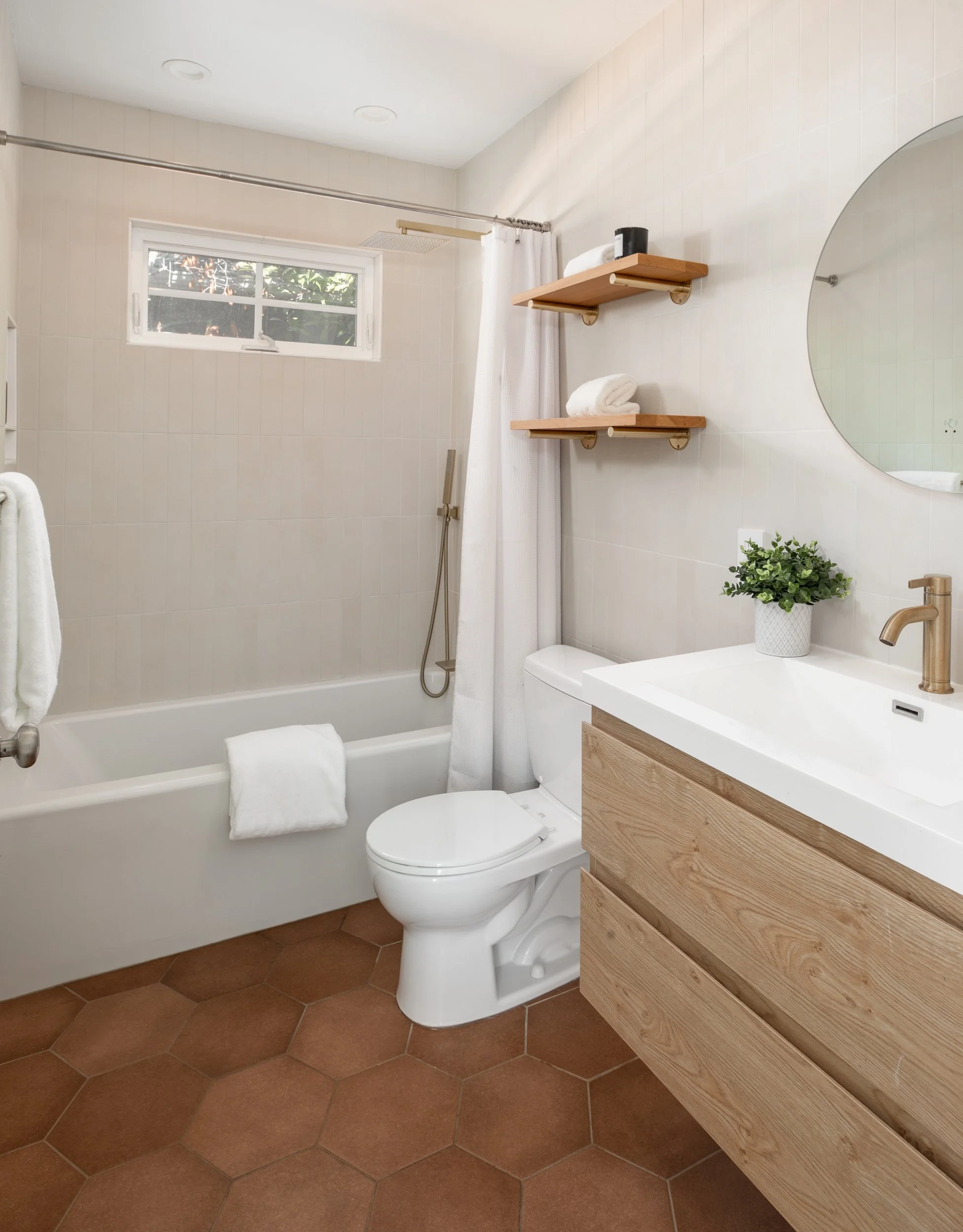 Bathroom with white tiled walls, a window with a view of greenery, a shower with a white curtain, a toilet, a wooden vanity with a white sink, a round mirror, and decorative plant.