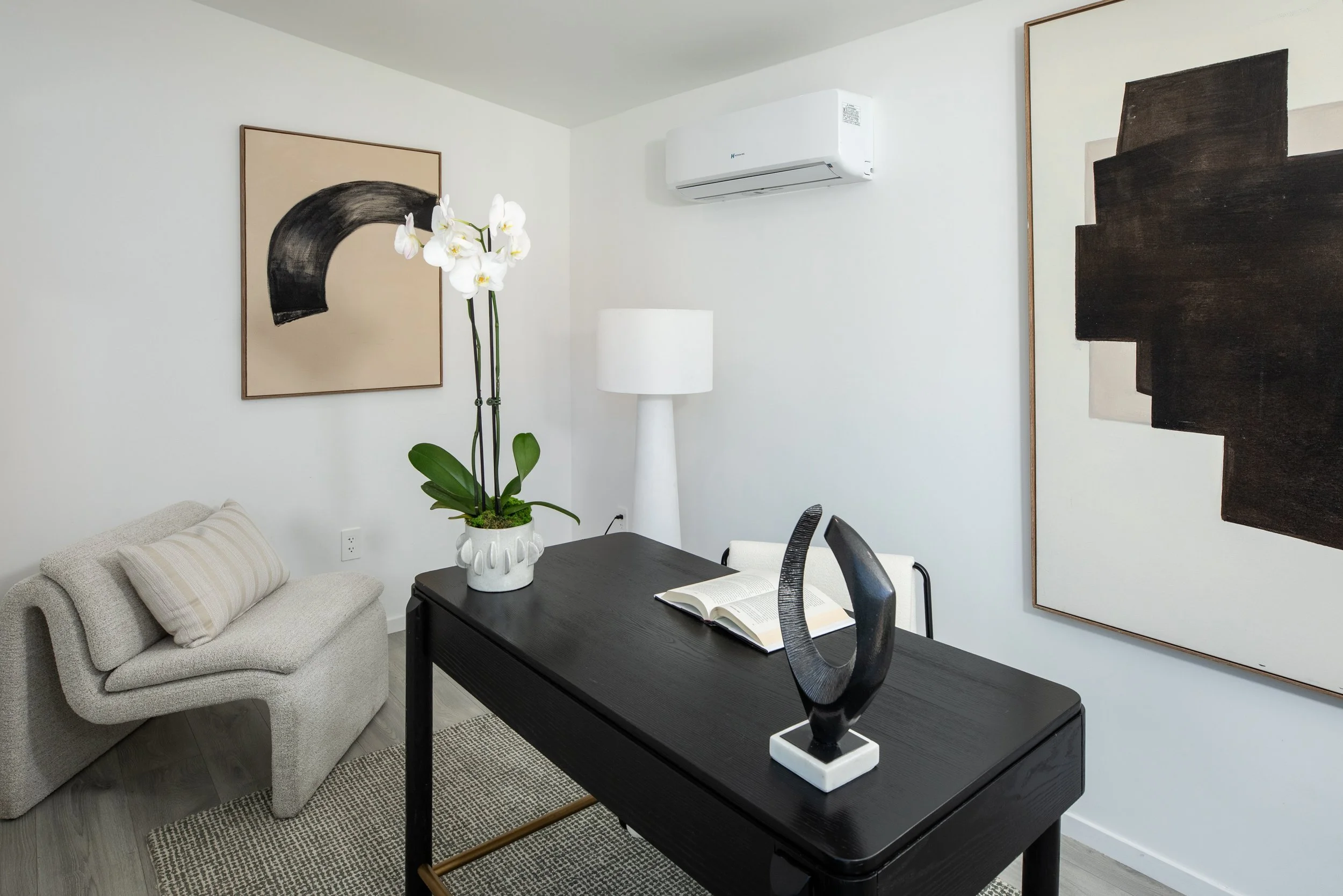 Living room with a beige sofa, a black dining table, a white orchid plant, paintings on the white walls, and a white air conditioner.