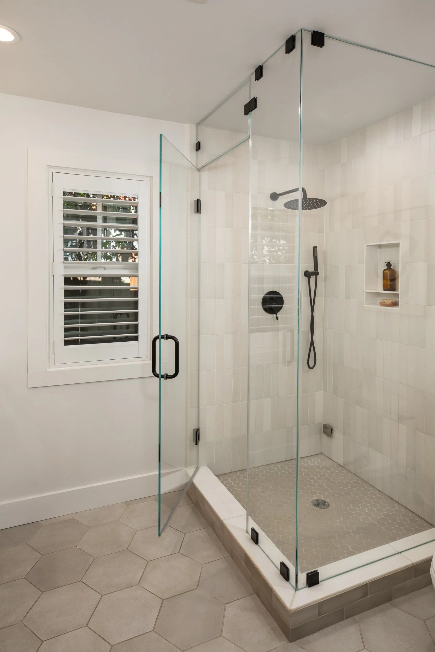 A modern bathroom shower with clear glass walls, black fixtures, a showerhead, and a built-in wall niche holding a bottle and a soap dish. The shower has a small tiled floor and is adjacent to a white wall with a louvered window.