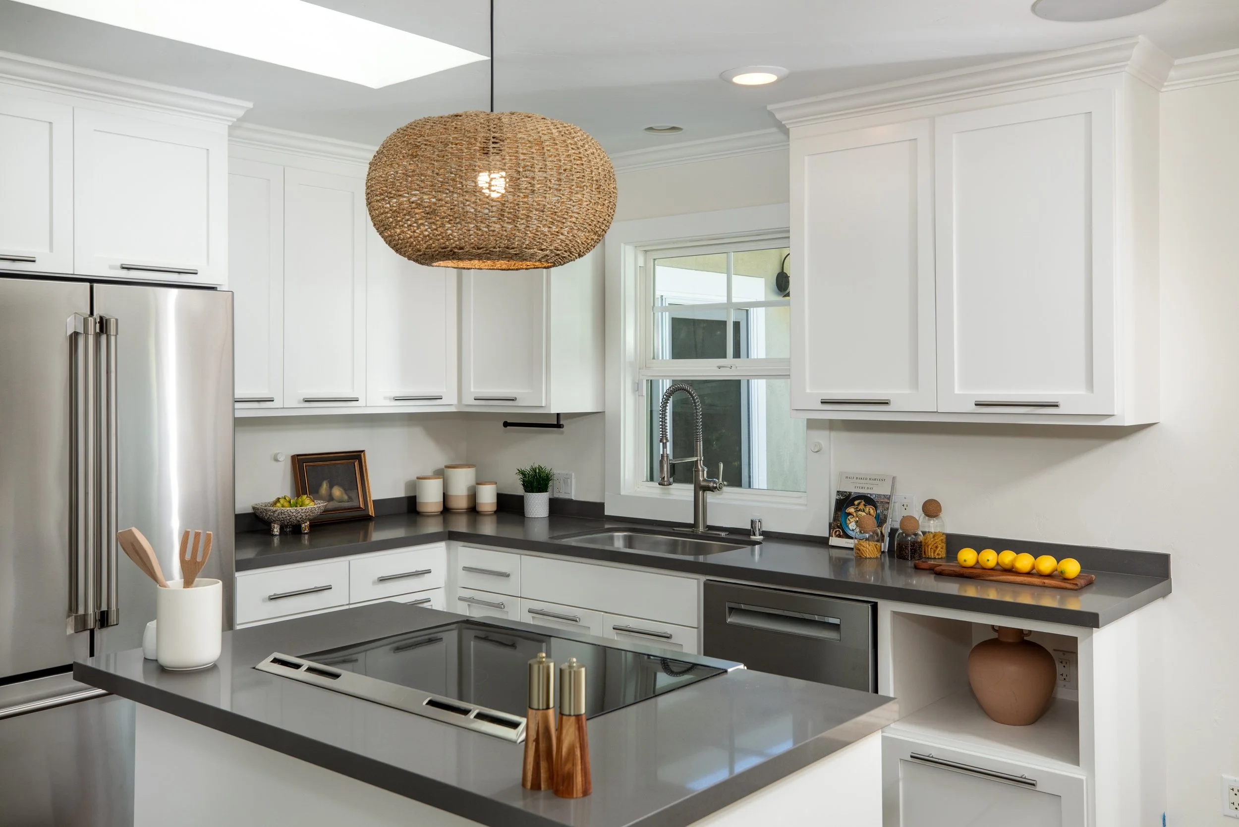 A modern kitchen with white cabinets, a stainless steel refrigerator, a black countertop, and a gray kitchen island with a sink and faucet. There is a woven pendant light hanging from the ceiling, a window above the sink, and various decorative items
