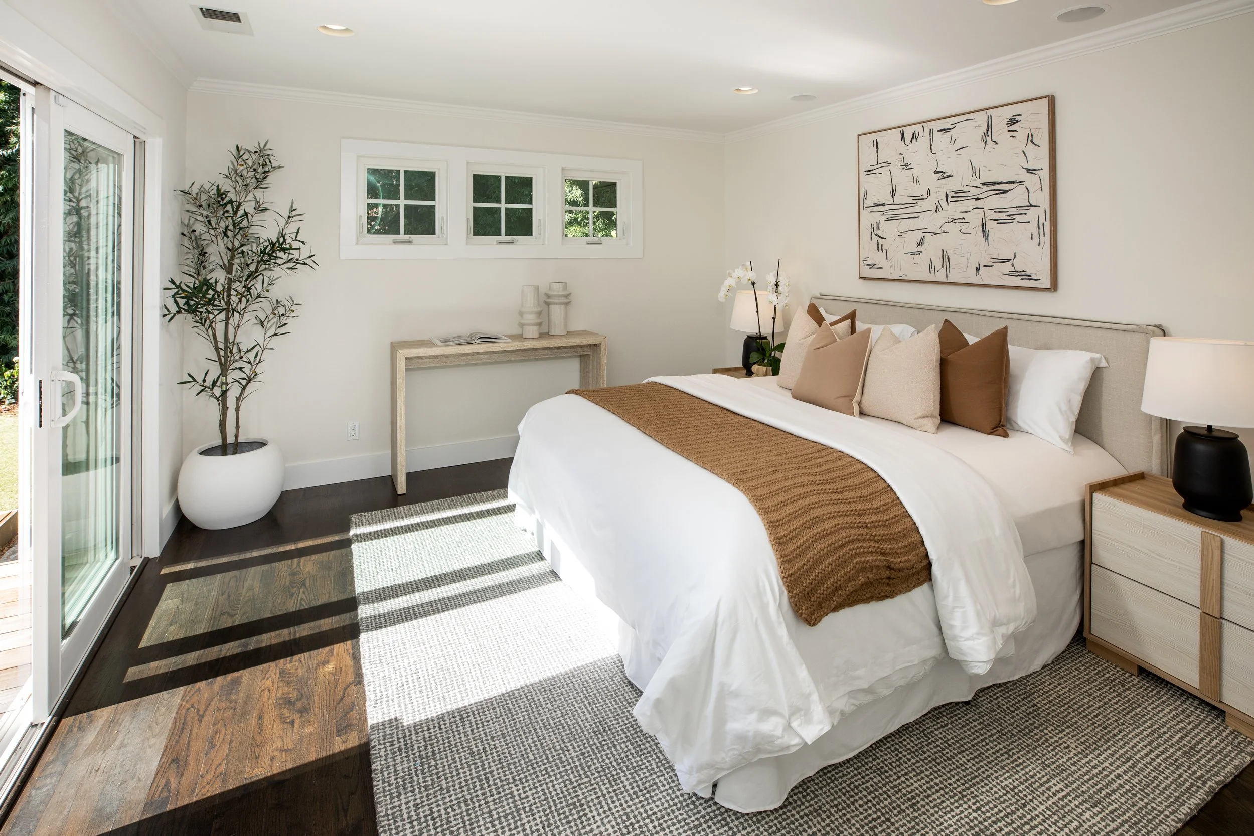 A bright modern bedroom with white walls and dark wood flooring. It features a large bed with white linens, beige and brown pillows, and a brown throw blanket. There is a nightstand with a black lamp and a plant with white flowers. A large abstract b