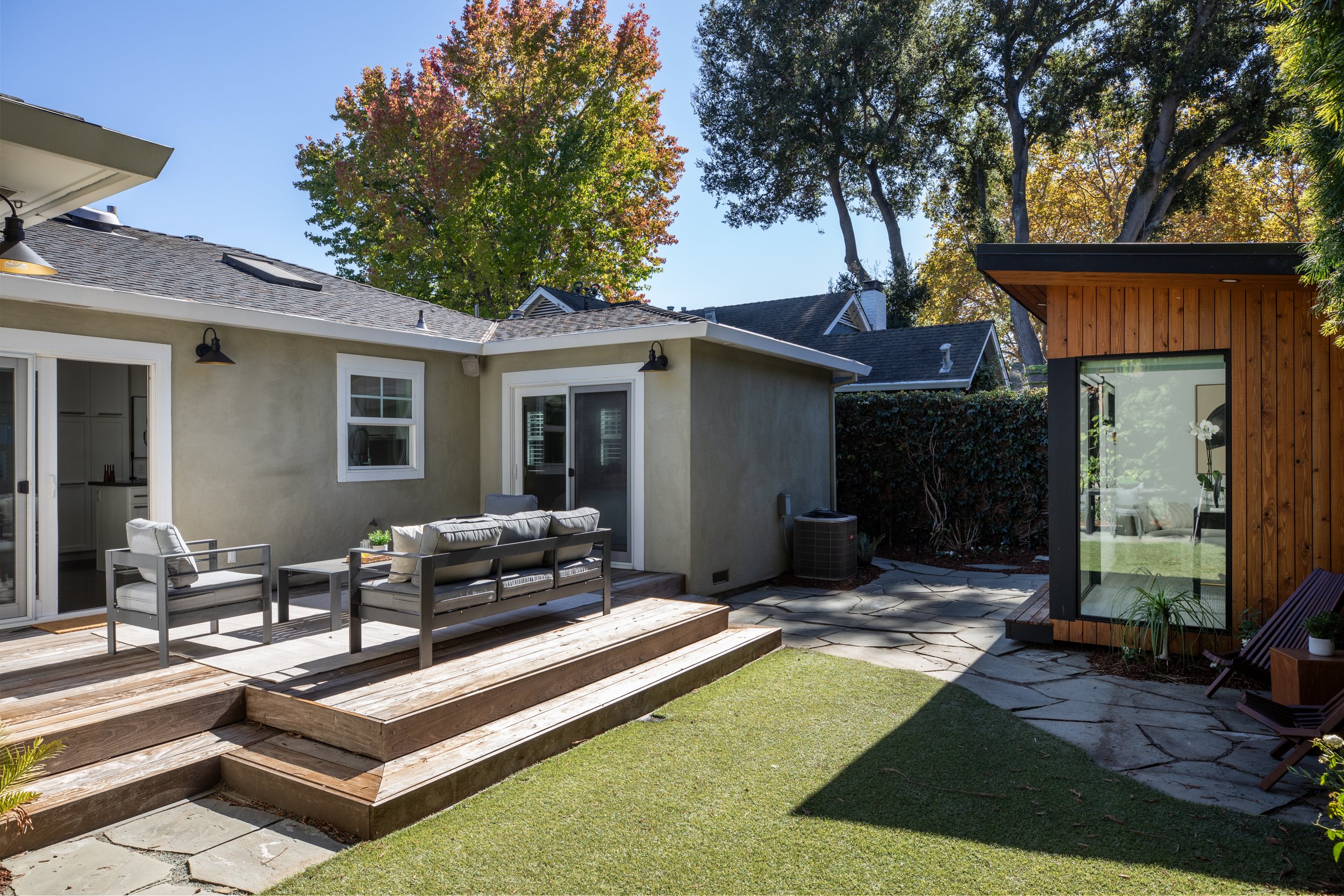 A backyard patio with outdoor seating, wooden deck, stepping stones, and a garden area. There are trees in the background and parts of houses visible.