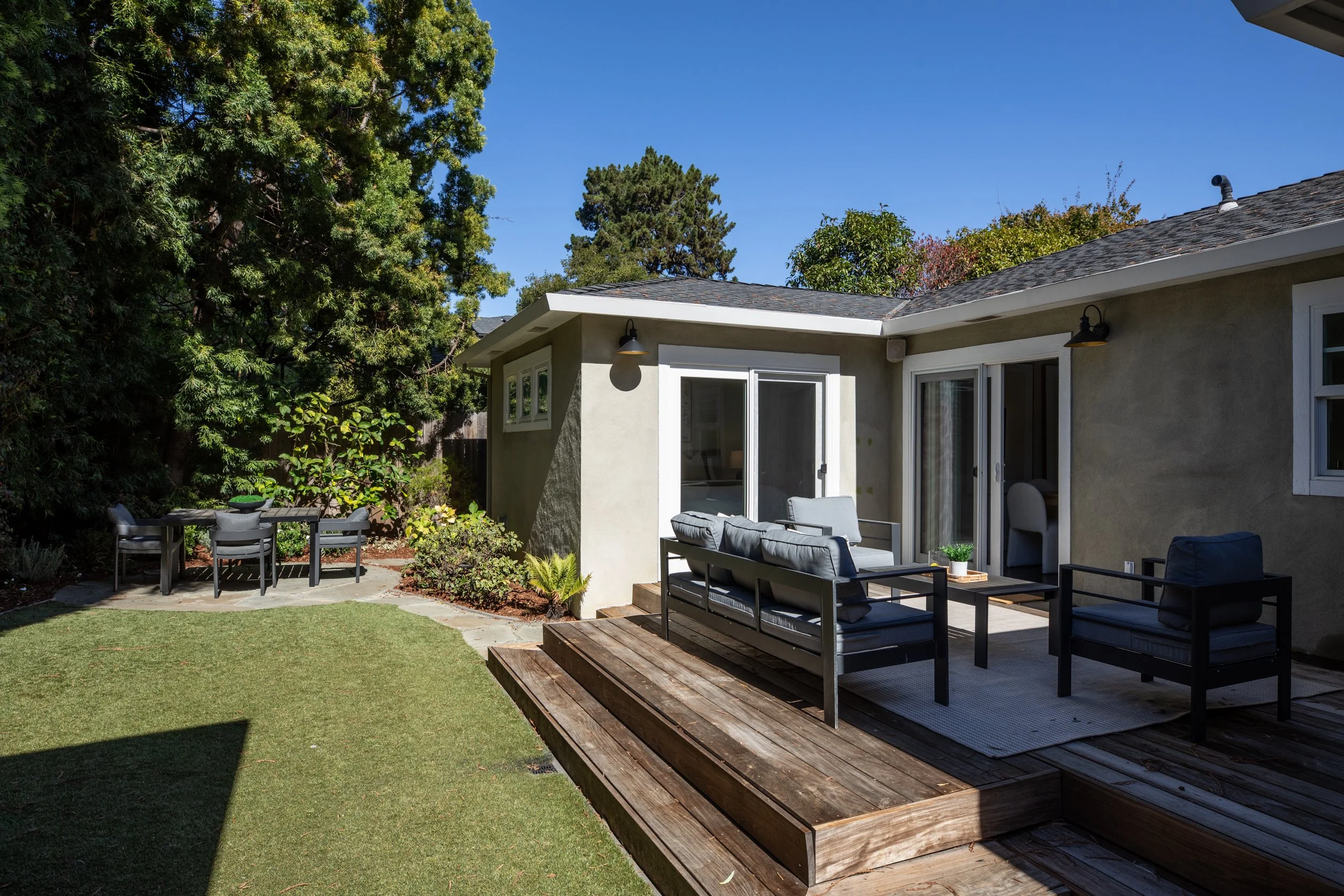 Back patio of a house with seating area and small table, a wooden deck, surrounding garden grass, tall trees, and a clear blue sky.
