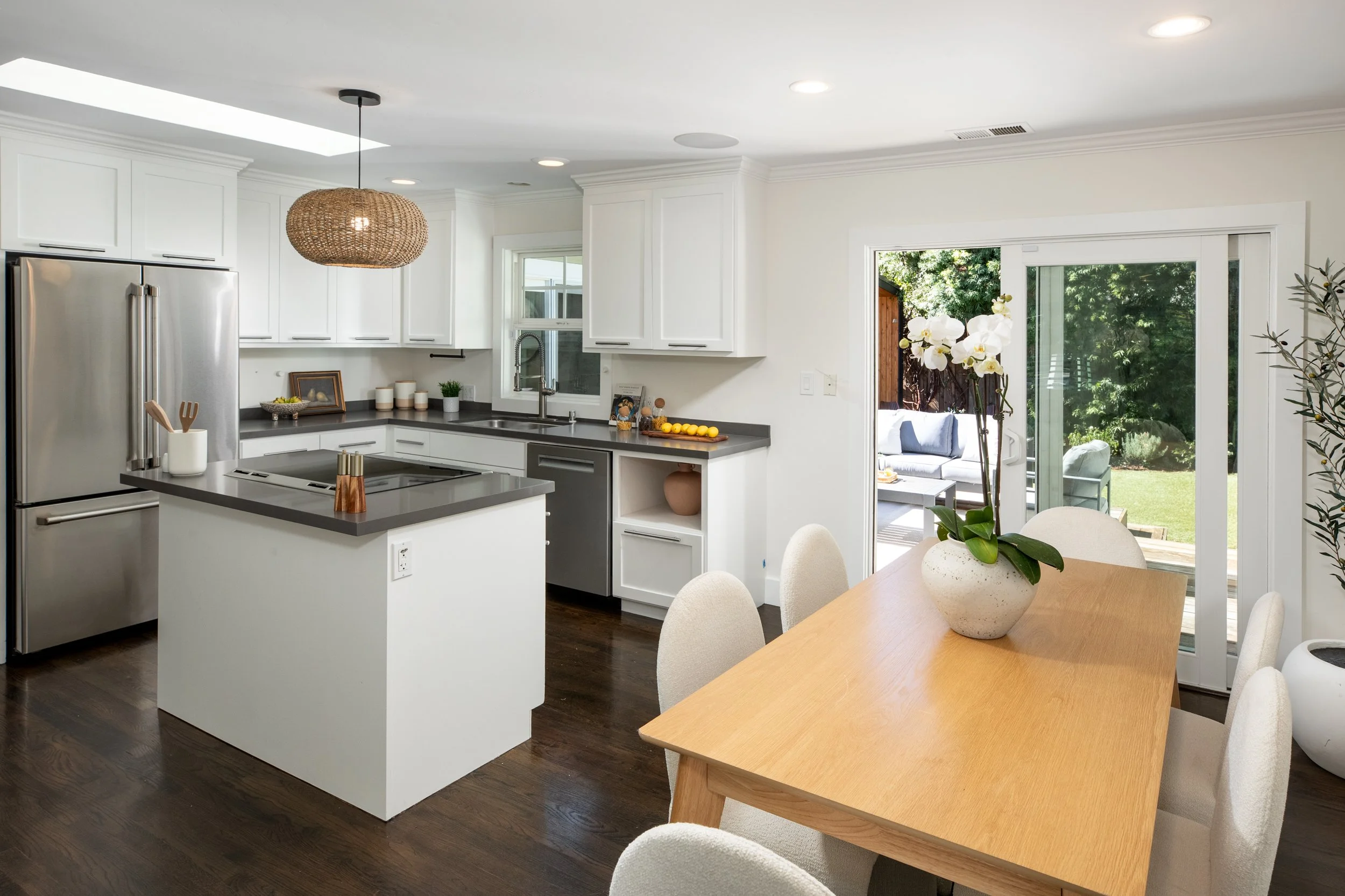 A modern kitchen with white cabinets, stainless steel refrigerator, a gray kitchen island with a utensil holder, a window above the sink, and a sliding glass door leading to an outdoor seating area with greenery.