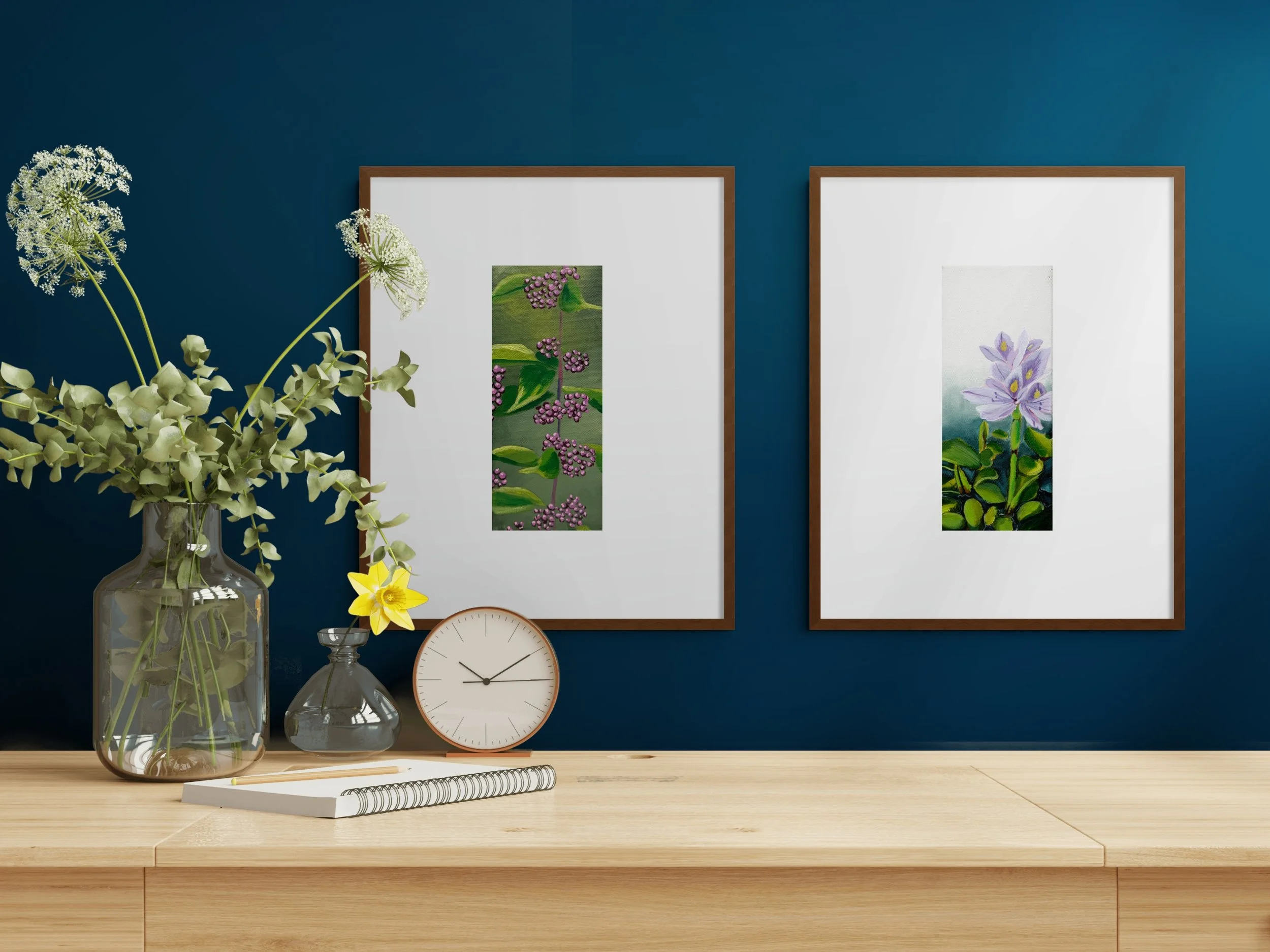 Blue wall with framed botanical artwork, a vase of greenery, glass decor, clock, and notebook on a wooden console table