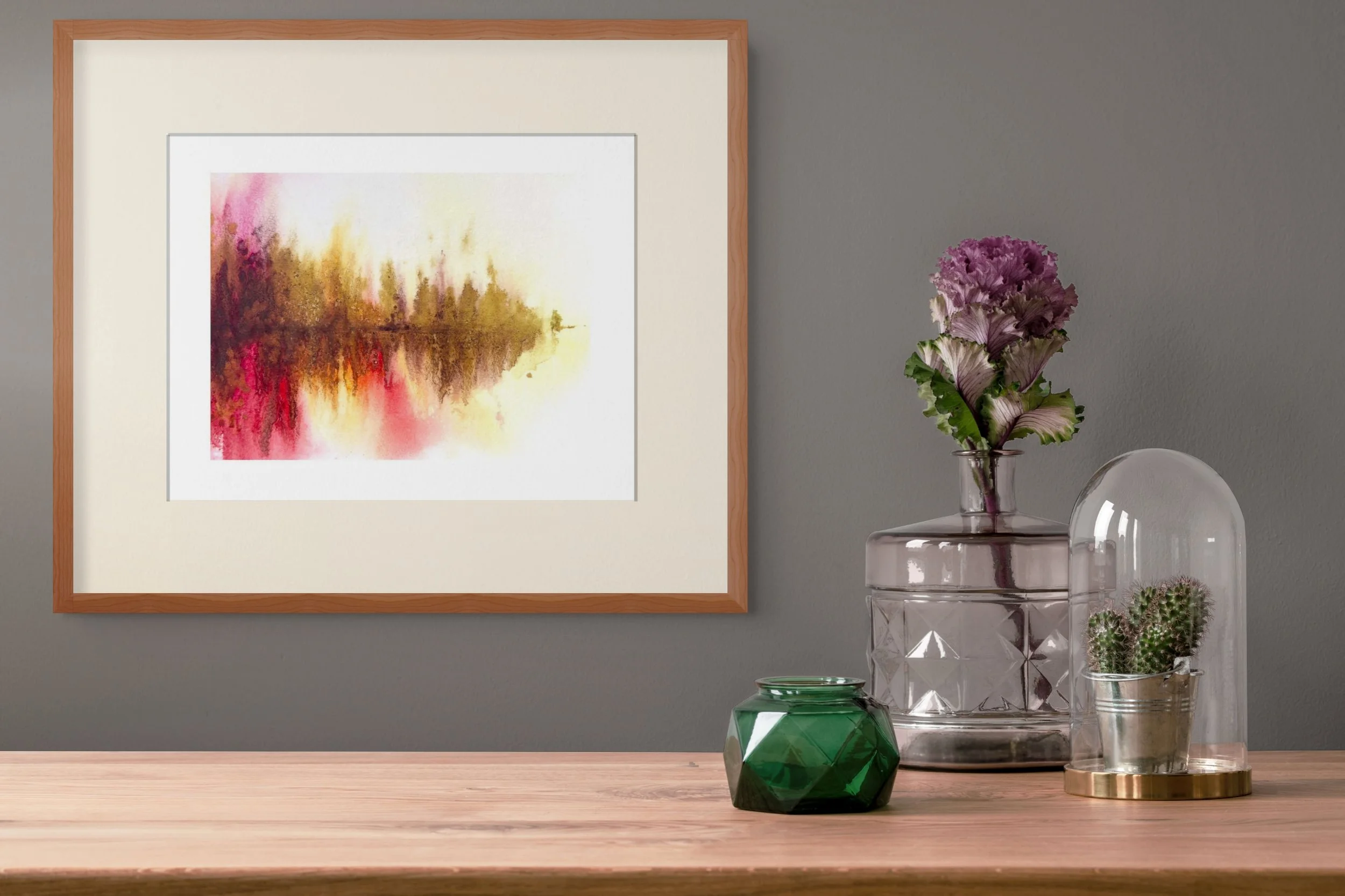 Framed abstract watercolor landscape above a wooden console decorated with a flower-filled glass vase, green glass jar, and cactus under a glass cloche.