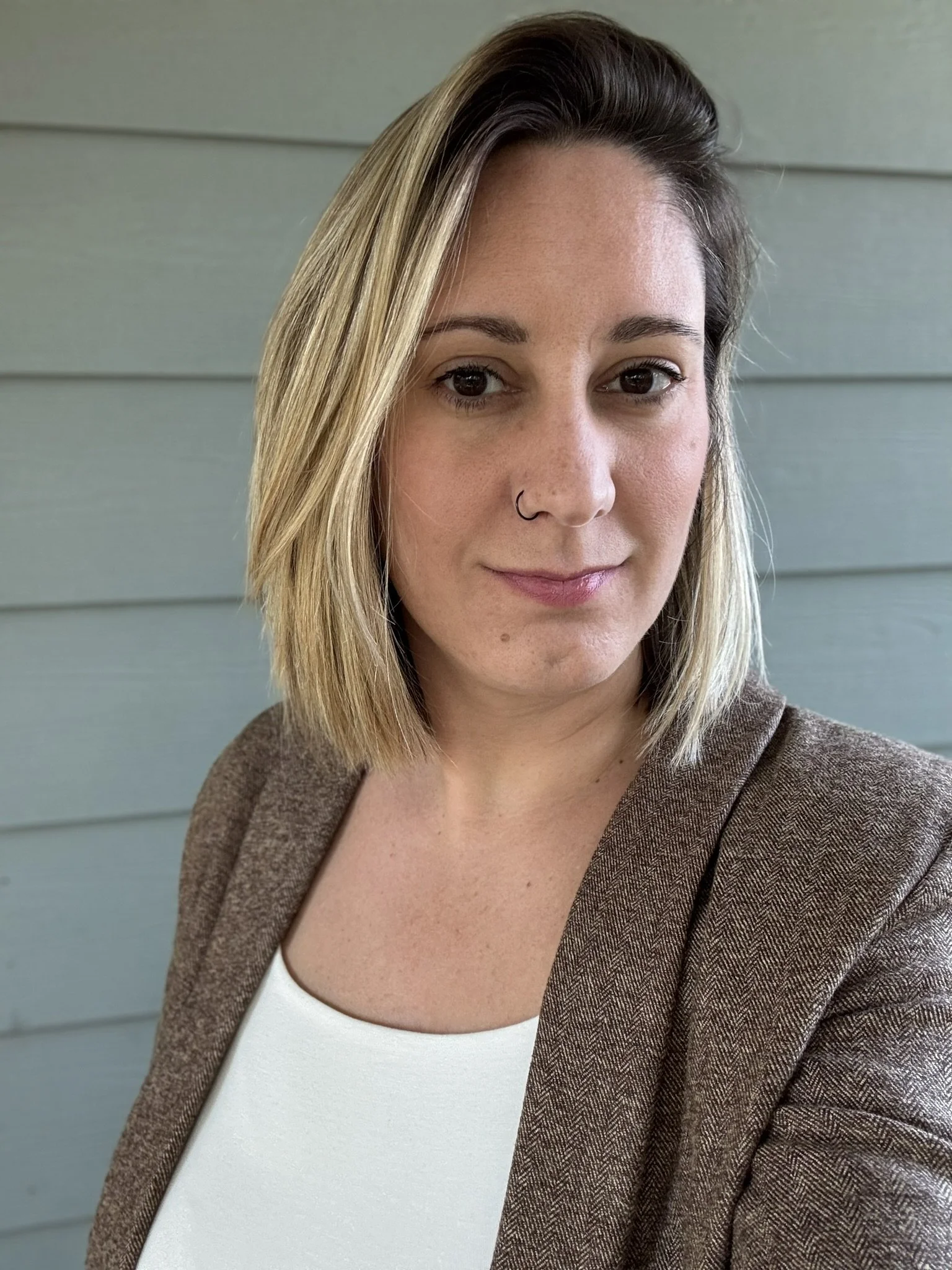 Portrait of a smiling woman with shoulder-length blonde hair and a nose ring wearing a brown blazer against a blue-gray wall