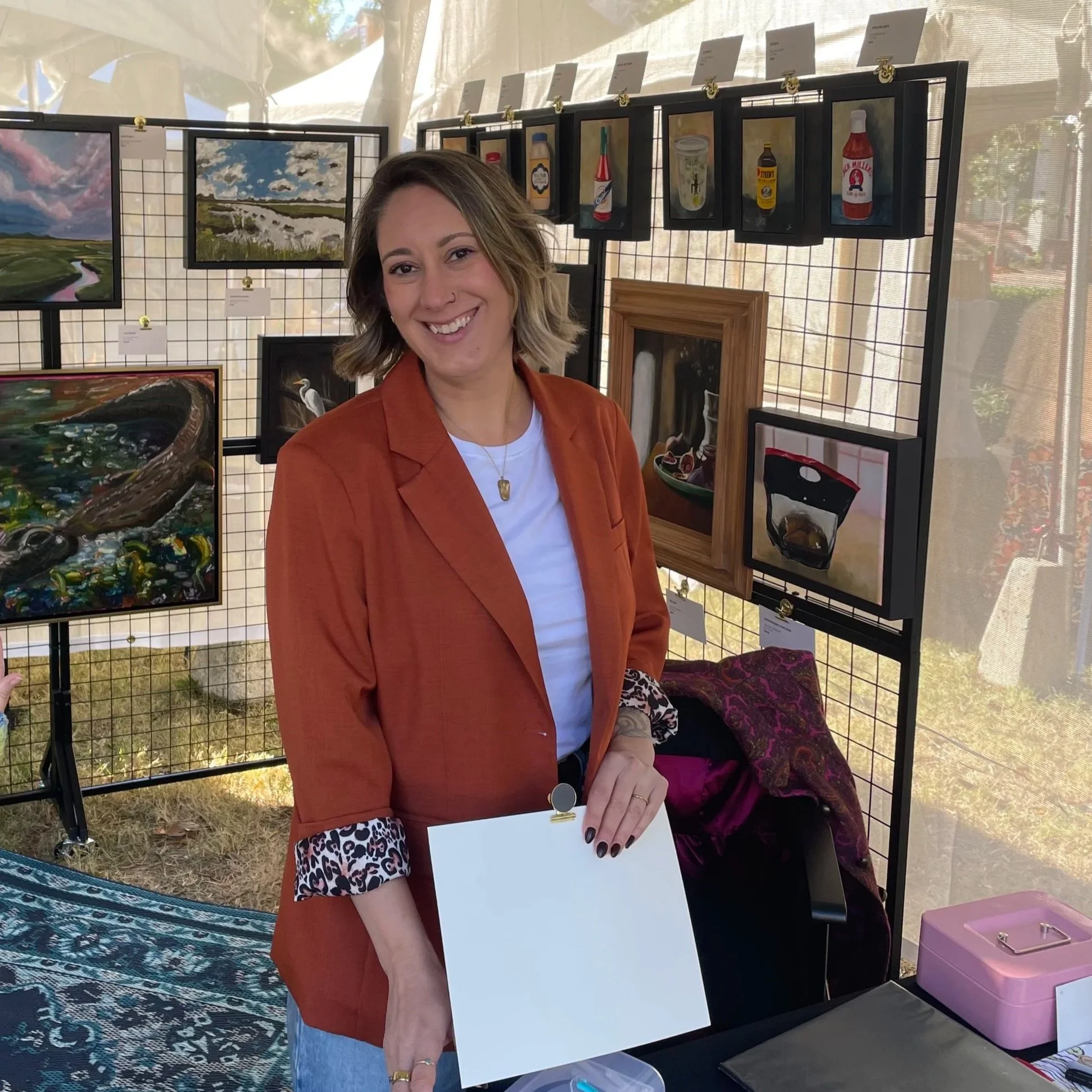 Smiling woman in an orange blazer holding a clipboard at an outdoor art exhibition displaying paintings and framed artwork.