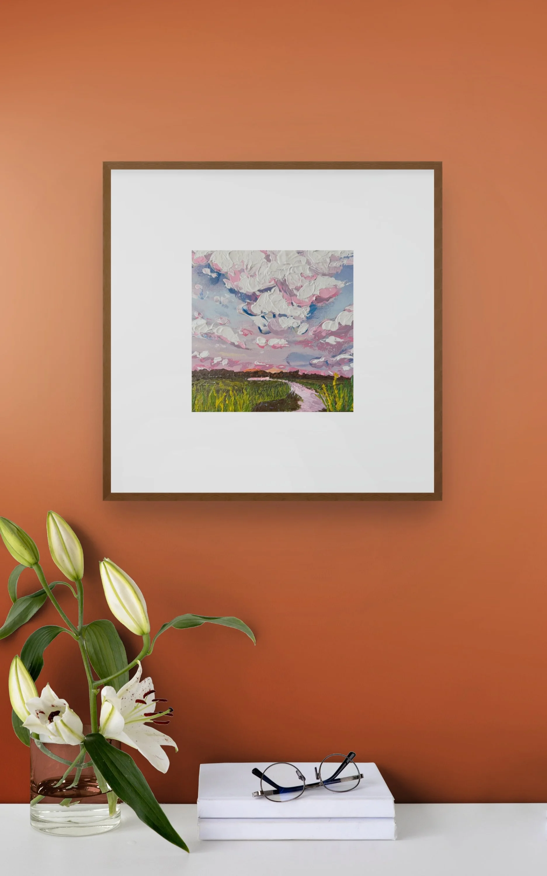 Framed impressionist landscape painting above white books and eyeglasses, with white lilies in a glass vase on an orange wall